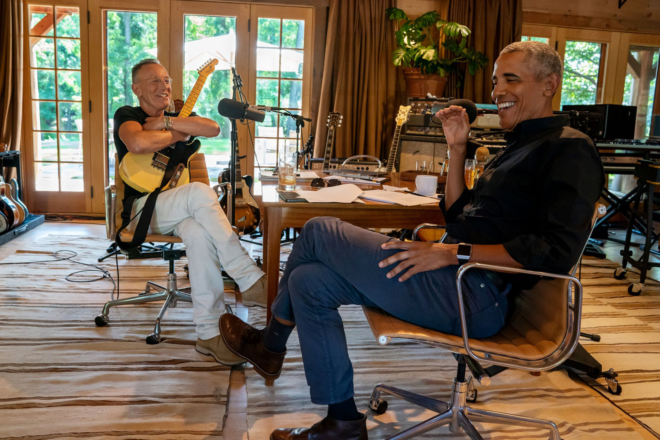 Bruce Springsteen, on the left, smiles while holding a guitar, and on the right Barack Obama laughs during a recording of their podcast