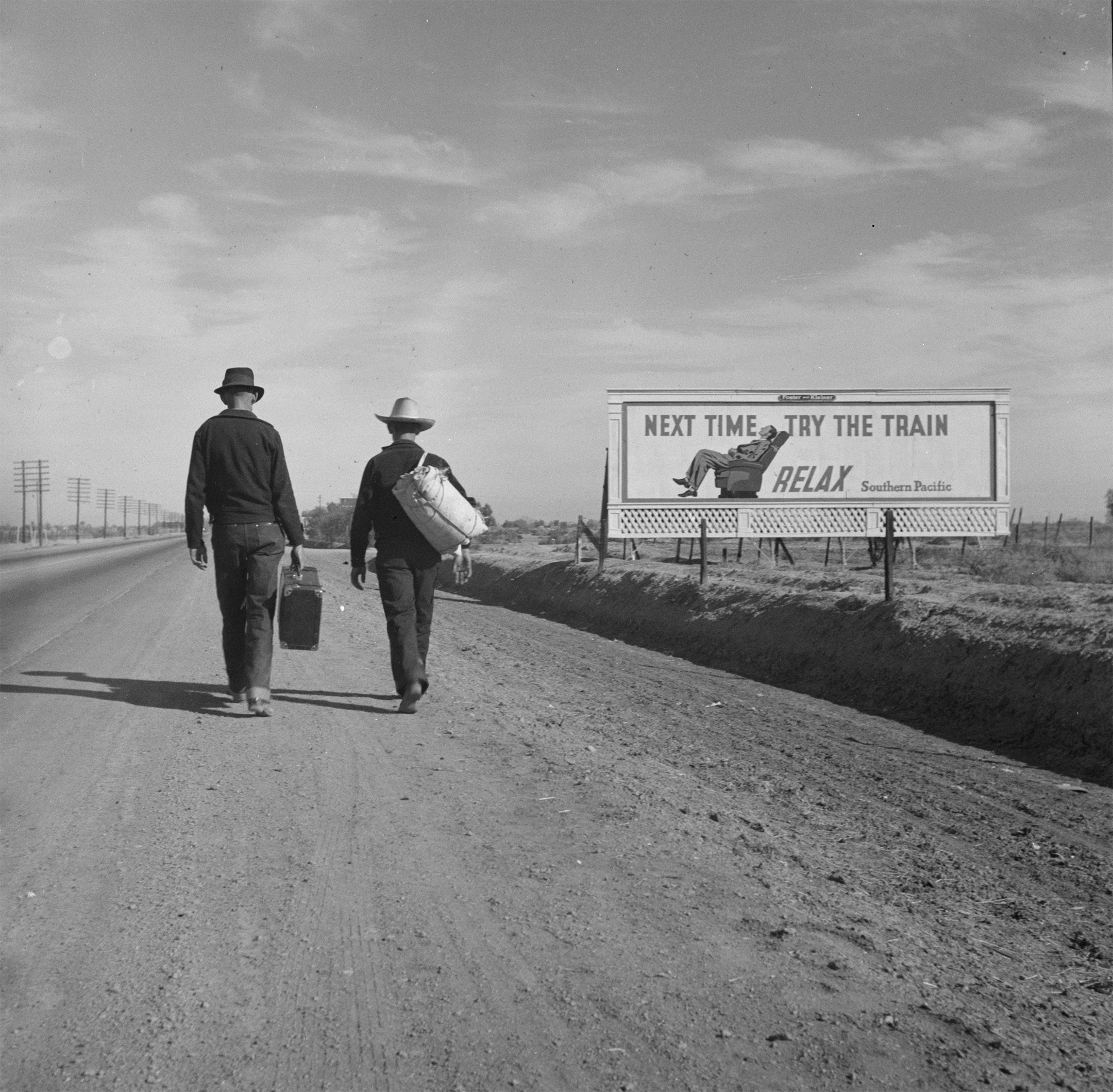 Black and white photo of two men, seen from behind, walking down a dusty road carrying bags and wearing cowboy hats, with a billboard to the right reading "next time try the train relax"