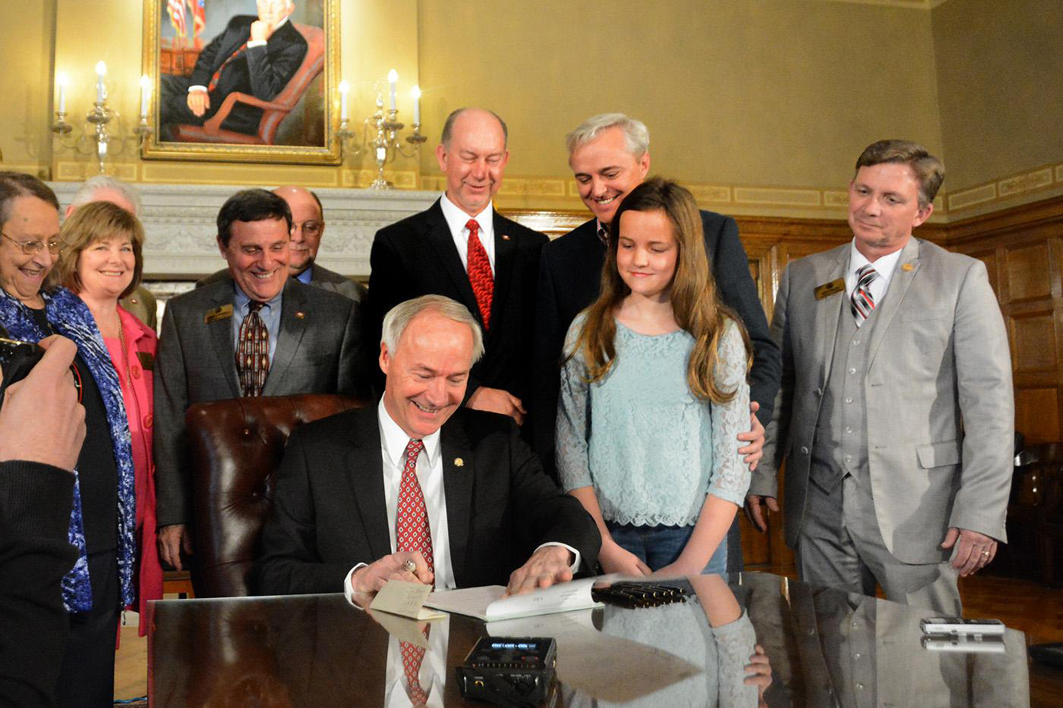 Asa Hutchinson smiles while seated at a table sighing legislation, surrounded by nine onlookers