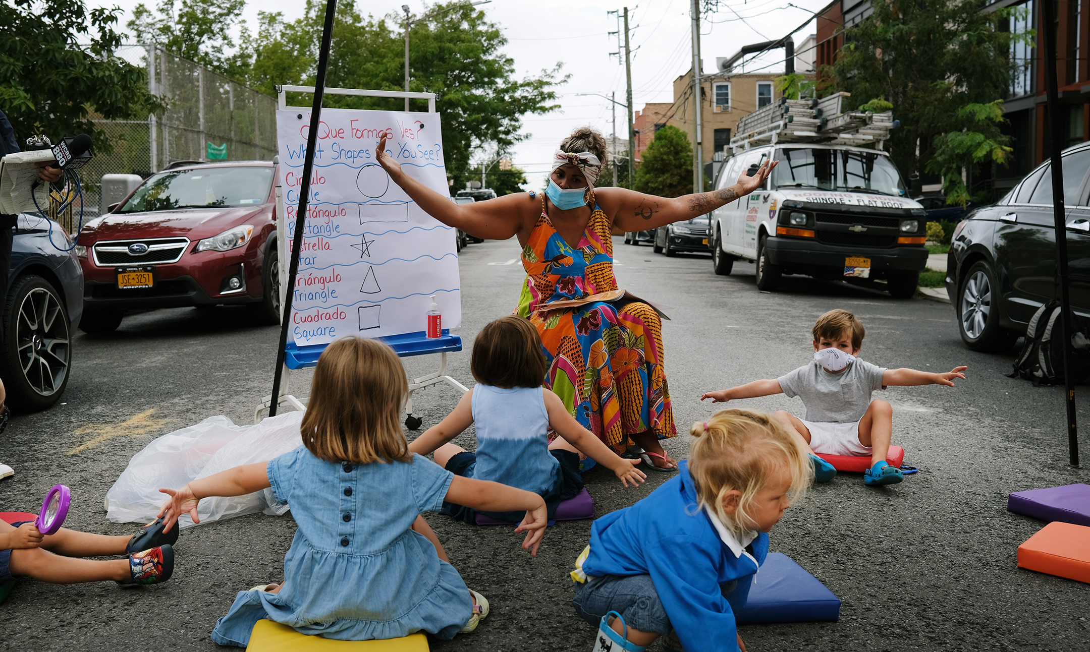 A teacher seated in front of six elementary school students conducts a lesson on the street in front of their school.