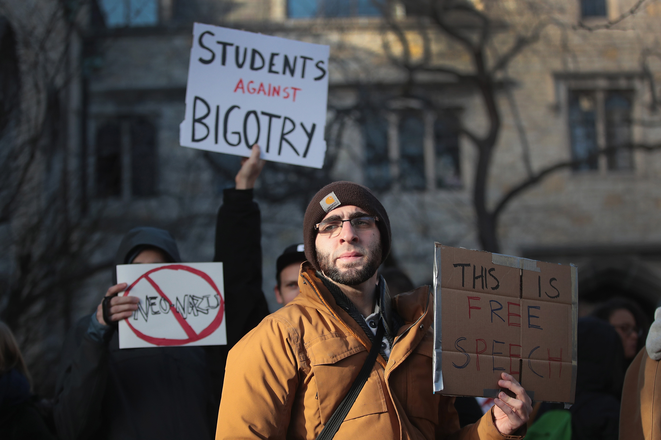 Man in a tan winter jacket and brown knit cap holds a sign made from a cardboard box that reads this is free speech
