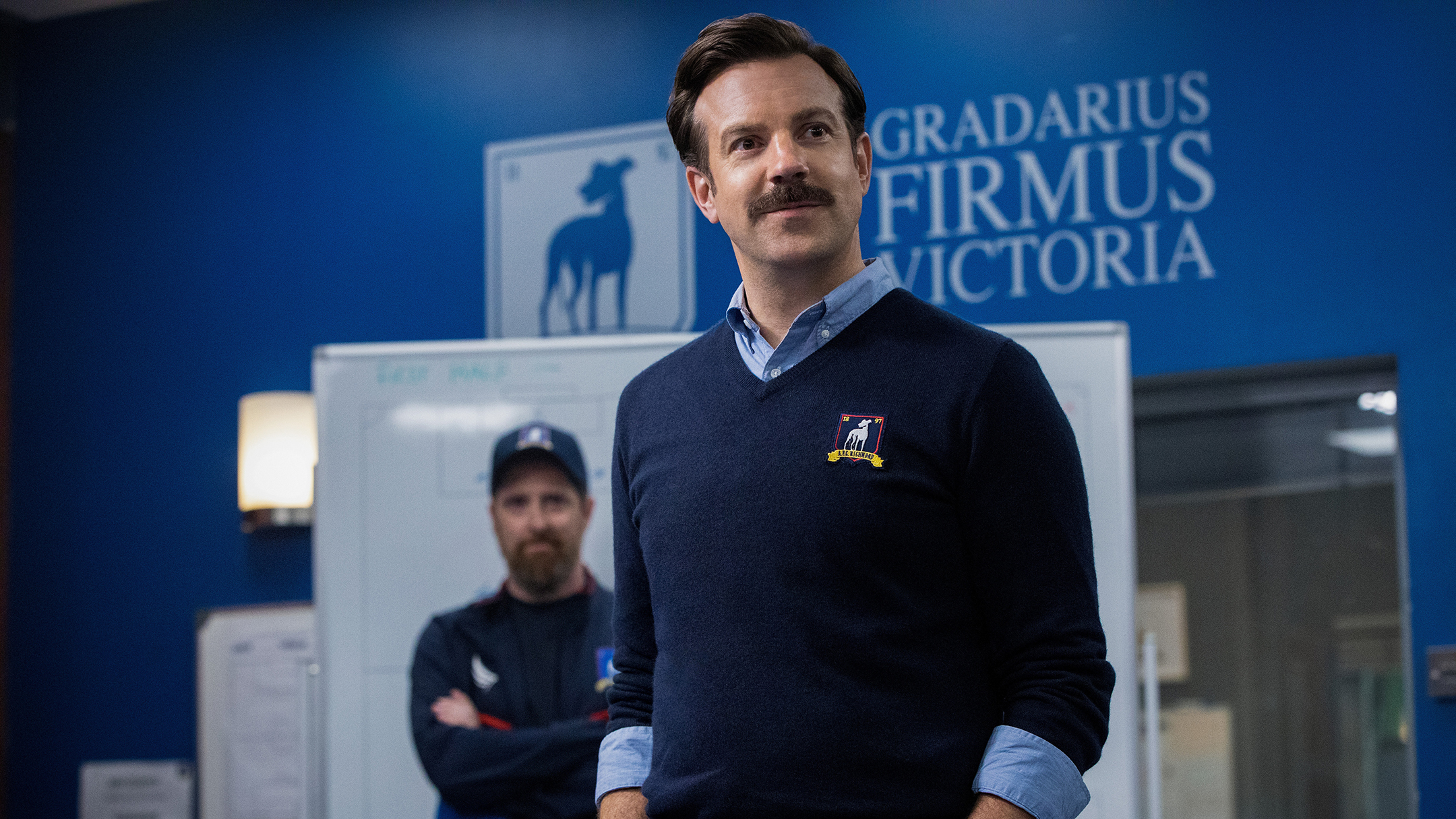 Photo from the show Ted Lasso showing a smiling Jason Sudeikis with his hands in his pockets standing in a locker room