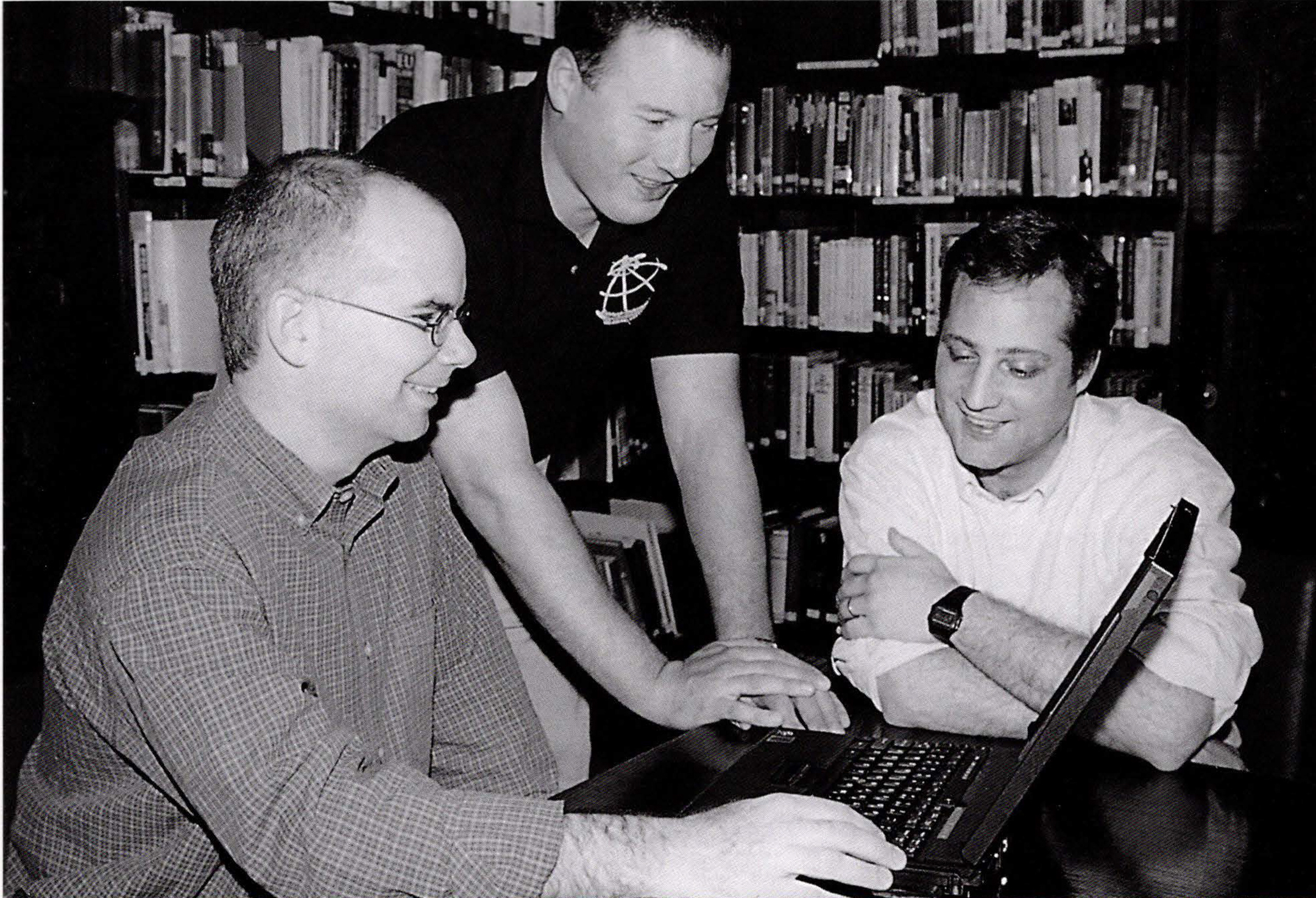 Black and white of three men smiling huddled around an open laptop