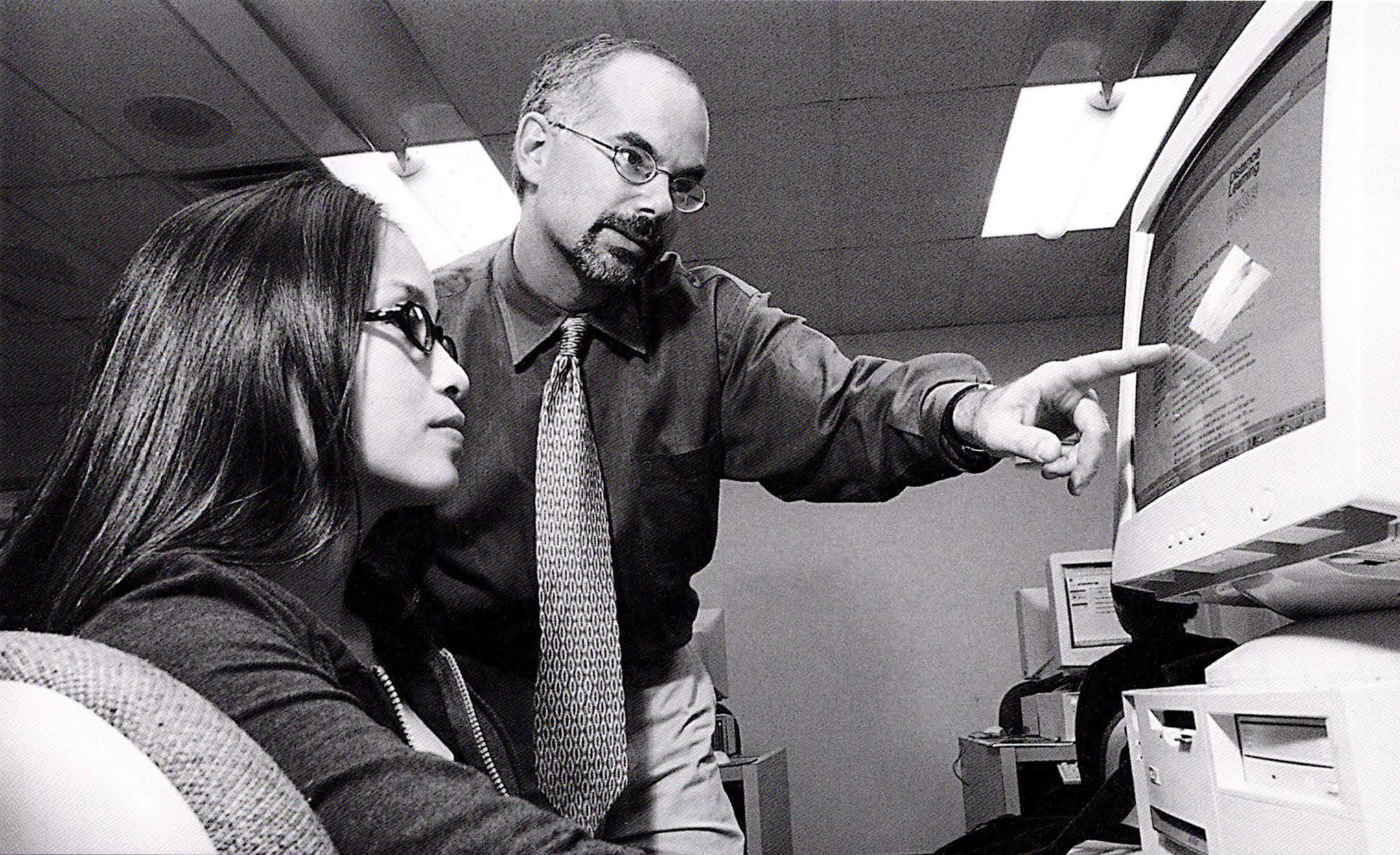 Black and white photo of a male teacher, standing, pointing at a computer monitor while a female student, seated, looks on