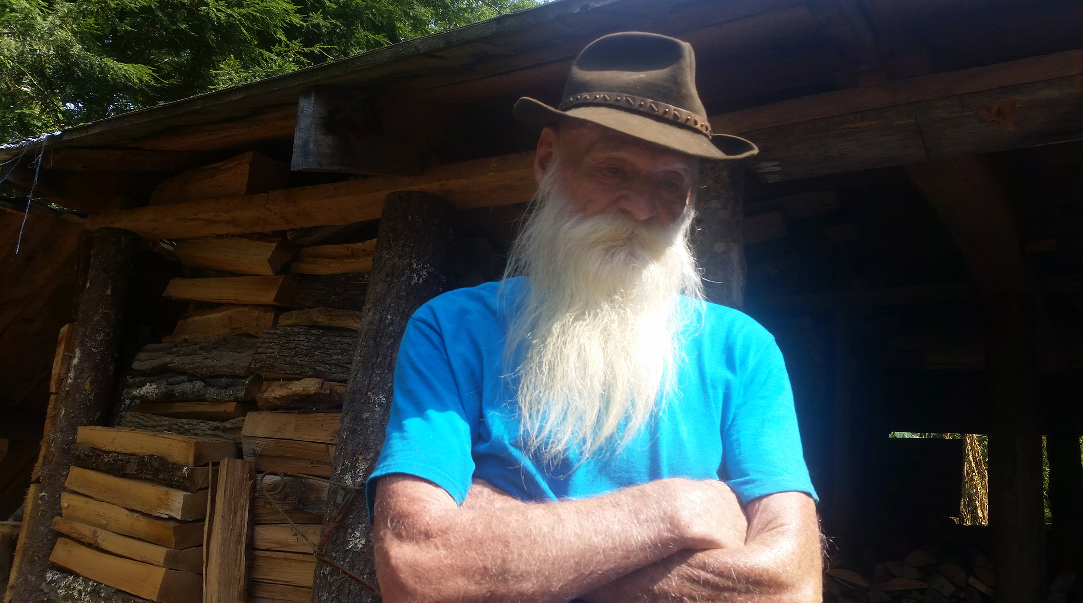 Old man with a long white beard in a blue t-shirt and brown hat stands in front of a cabin with his arms crossed and looking down