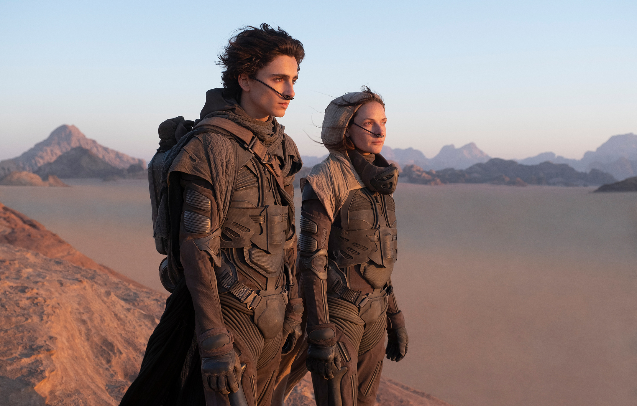 A young man and his mother, in dark futuristic clothes, stand in a desert and look to the right of the frame