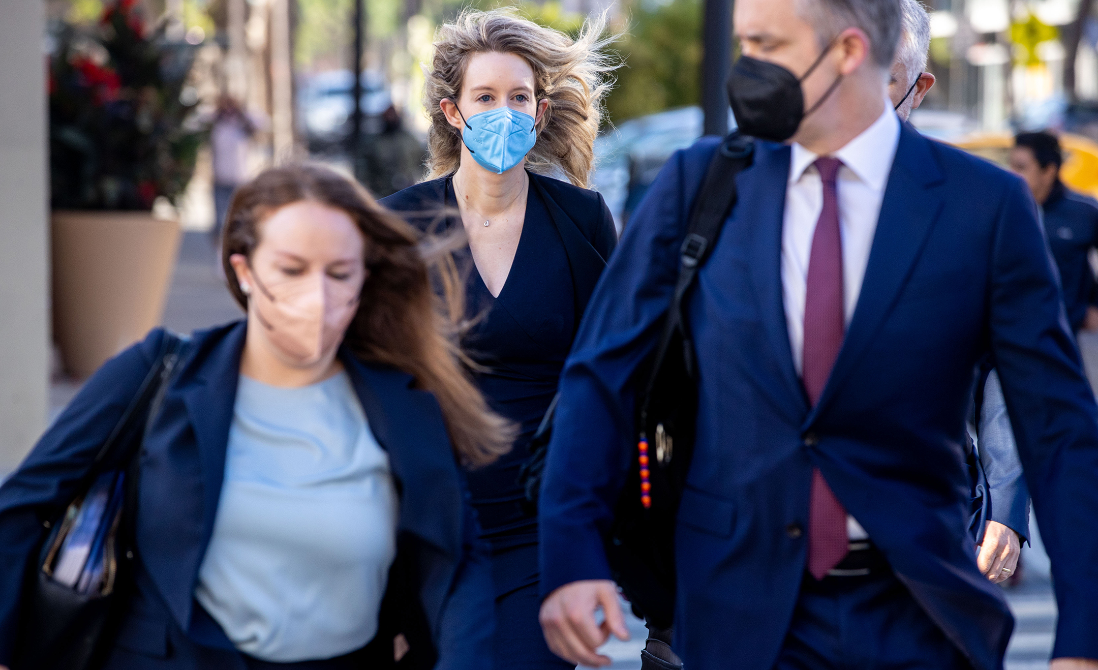 Tall blonde woman wearing a business suit and face mask walks behind two people, also masked, into a courtroom