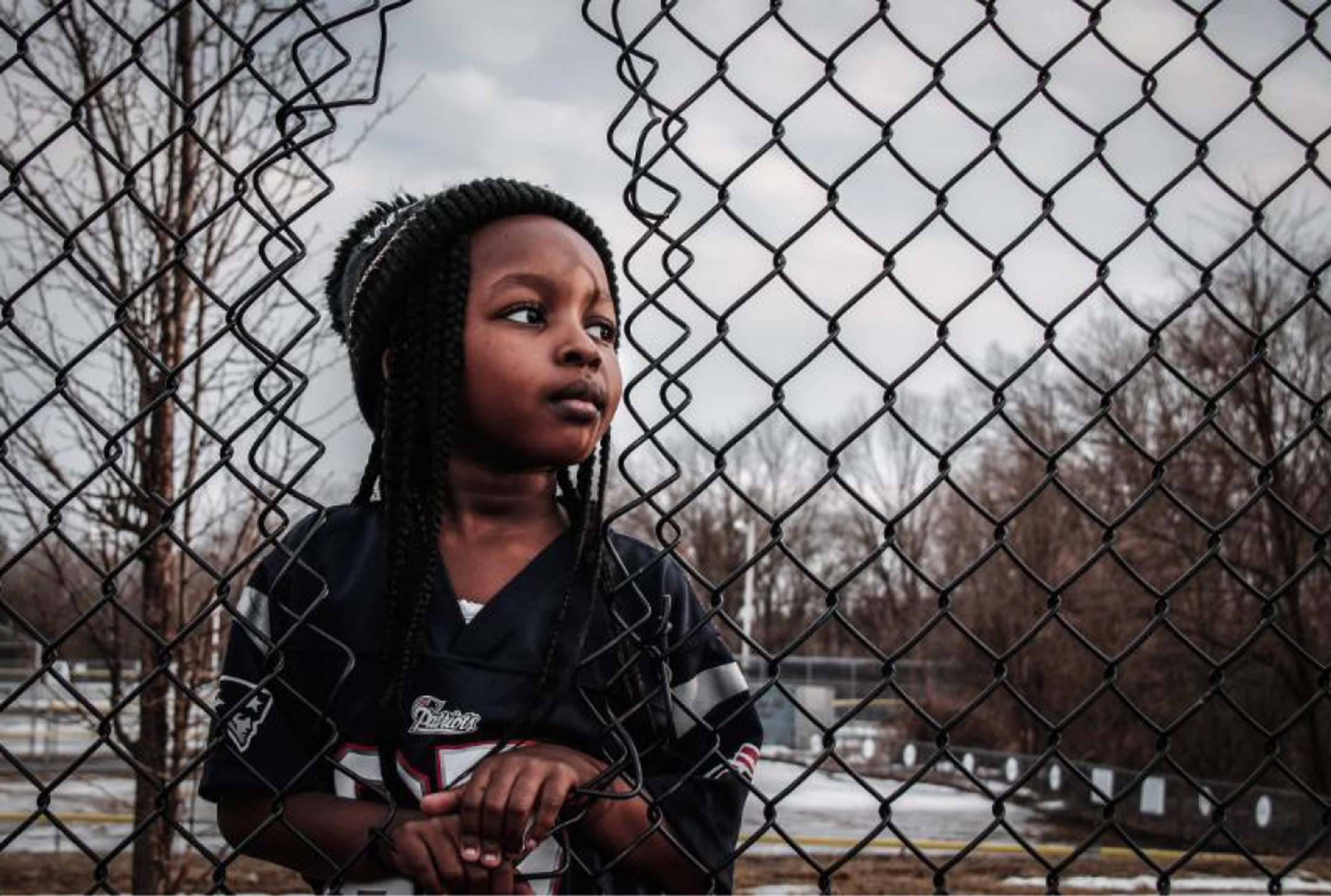 Photograph of a young black girl looking through a hole in a fence