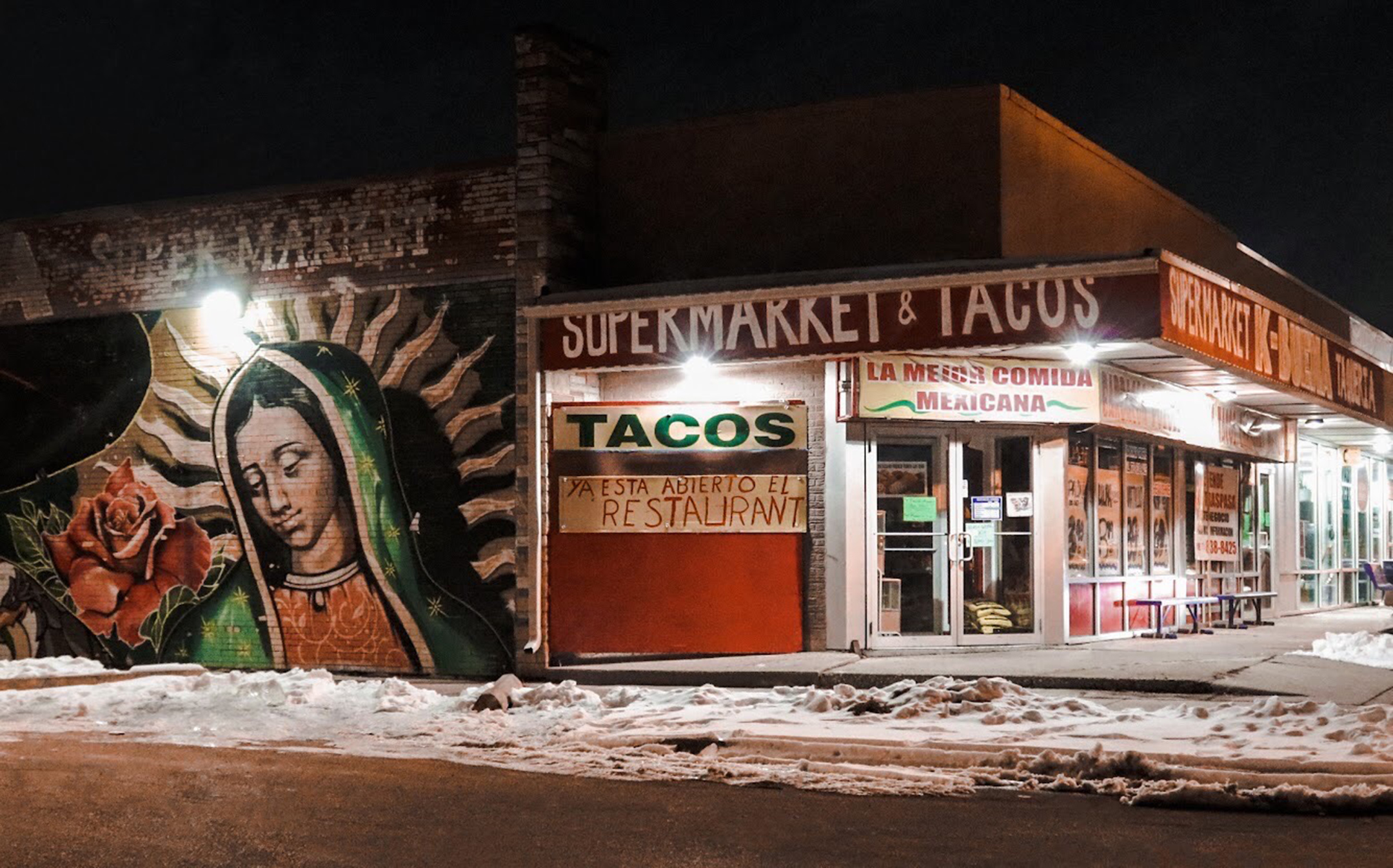 Nighttime photo of a supermarket and taco stand store front with a mural of the Virgin Mary on the left