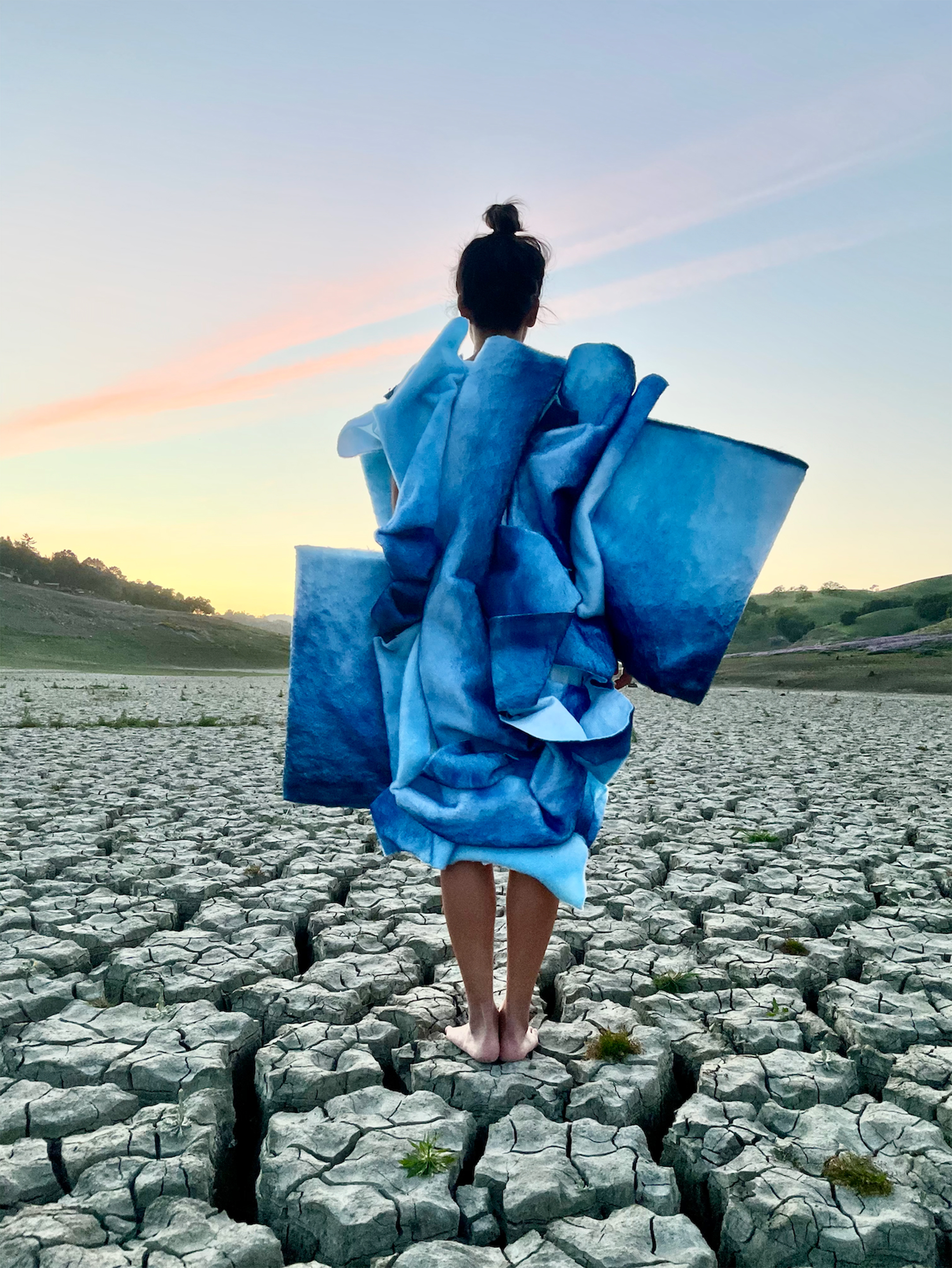 Photo of a woman, seen from behind, wearing a blue costume while standing on a parched ground at sunset