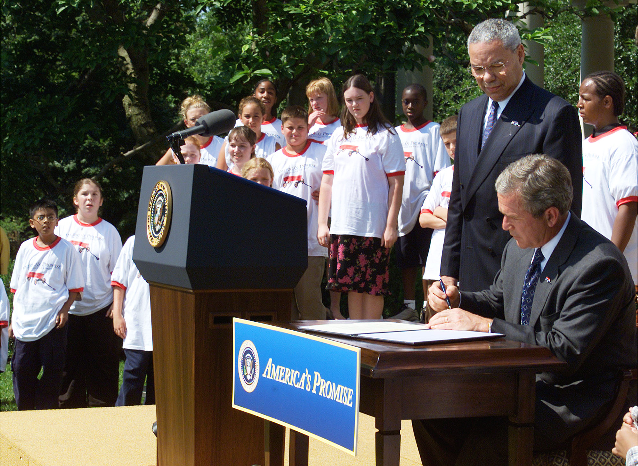Man sits at a desk while signing a document as another man and a group of high school students in matching white t-shirts look on