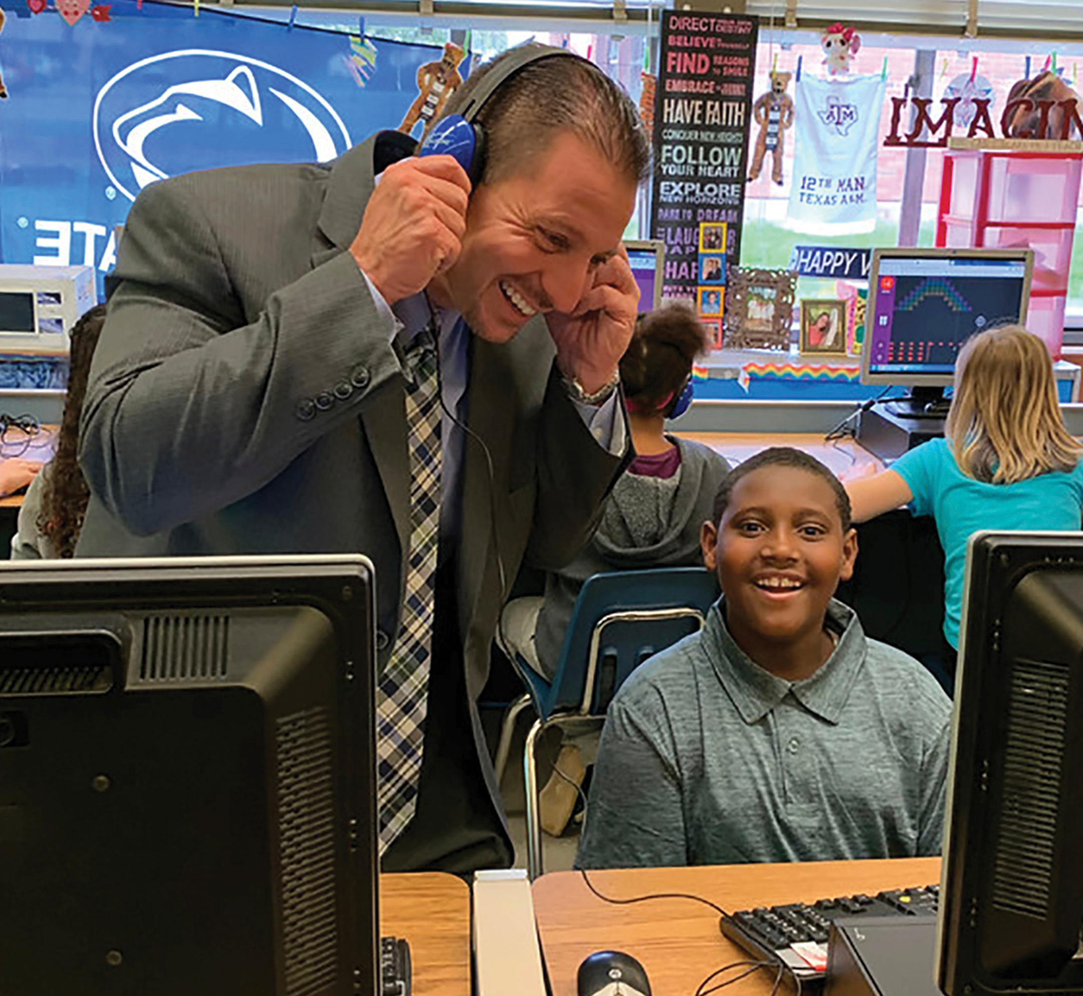 Standing male teacher wearing headphones smiles while working with a seated male student who is seated at a computer