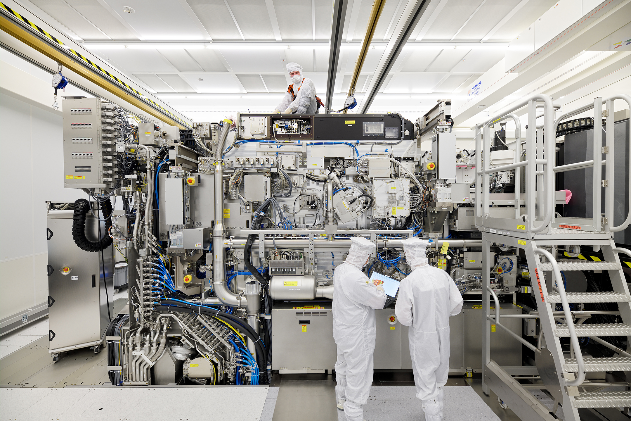 Three people in white clean-room body suits work on a machine in a white room