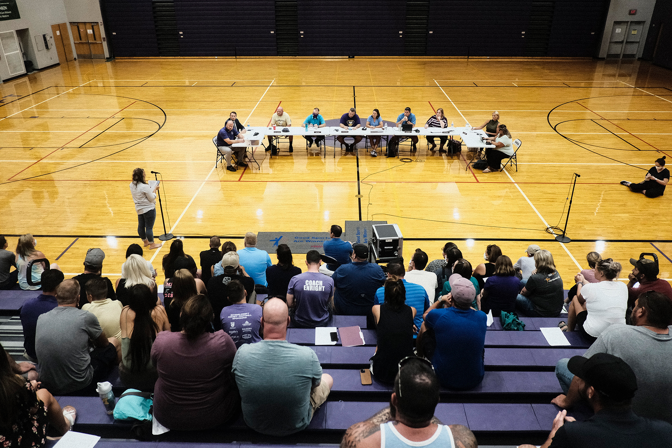 Photo from above of a row of people sit at tables in a gymnasium, in the back, a woman speaking at a microphone in the middle, and bleachers full of people listening in the front