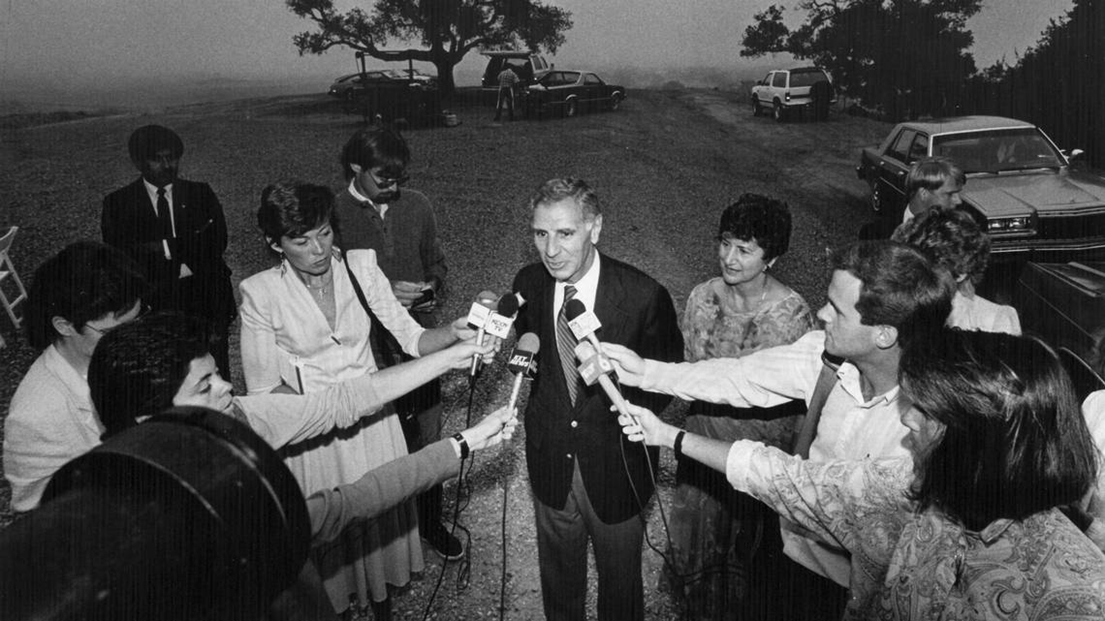Black and white photo of former California governor George Deukmejian standing in a scrum of reporters with microphones pointed at him