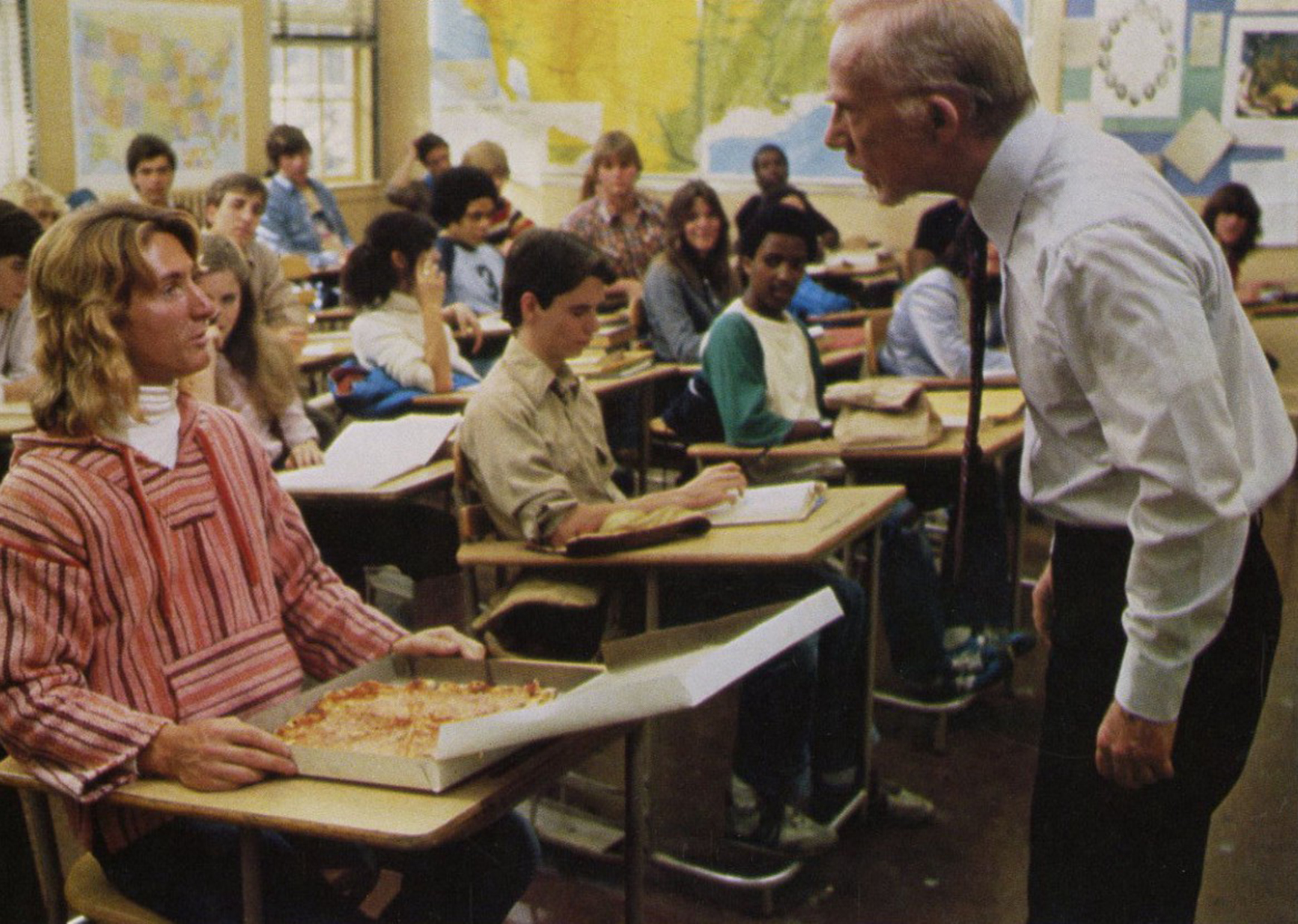 Movie still showing a student eating a pizza at his desk while being yelled at by his teacher