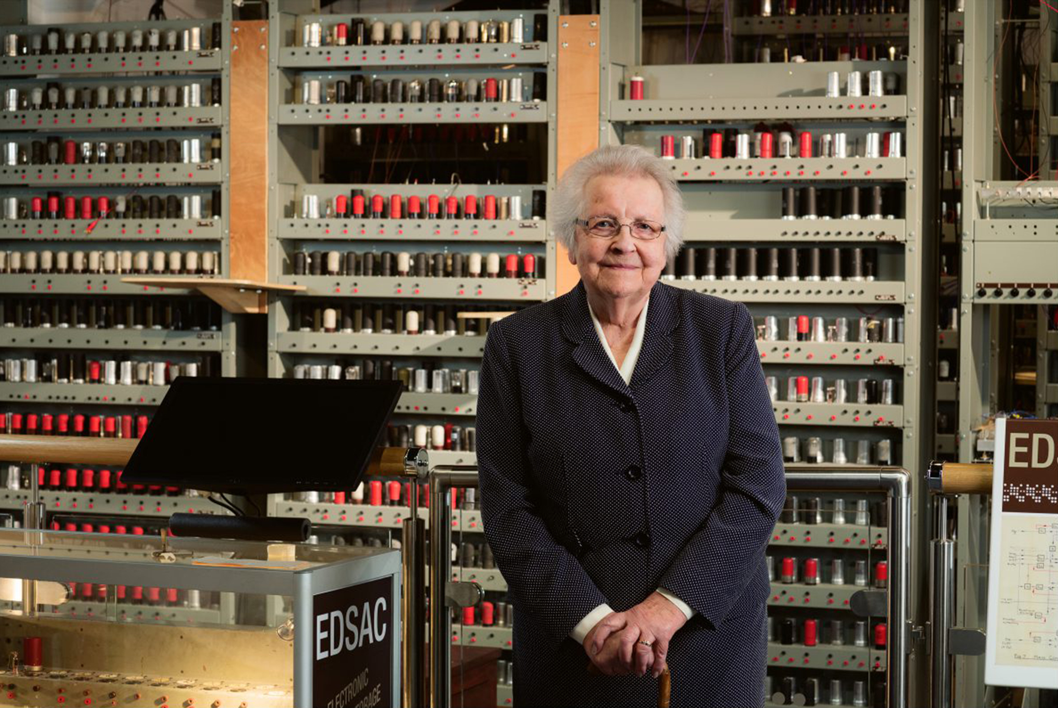 Older woman stands in front of a recreated EDSAC computer