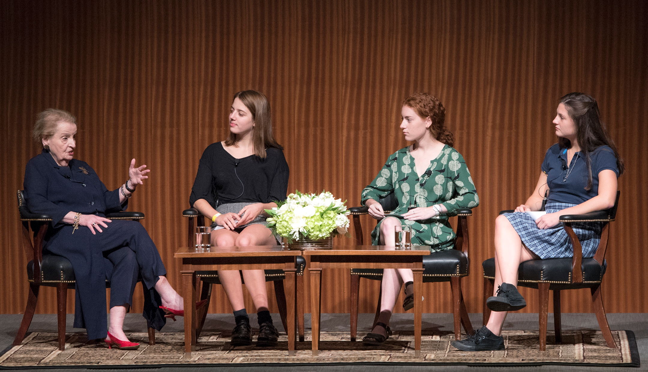 Madeleine Albright on a stage, at left, speaking with three female students