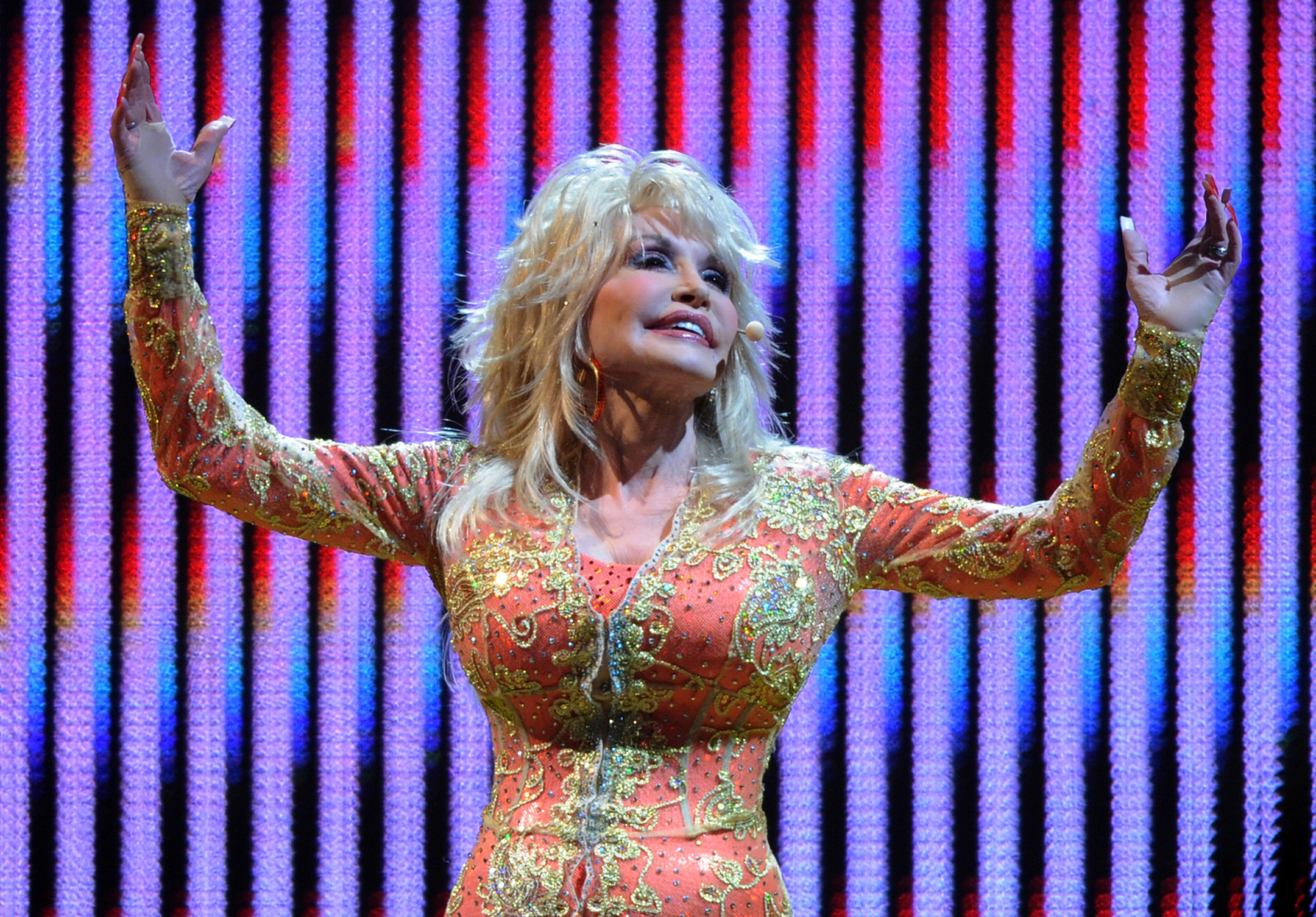 Photo of Dolly Parton, arms raised, against a multi-color background