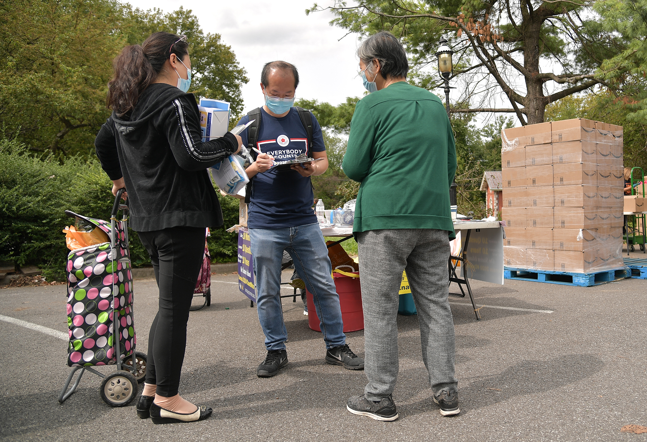 A census worker speaks to two people in a parking lot with a pallet of cardboard boxes in the background