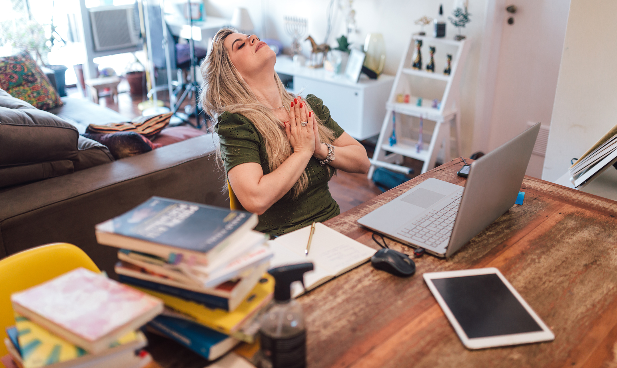 Woman sitting at desk in front of a computer, hands folded in a prayer pose, head back, eyes closed, in a cluttered home office