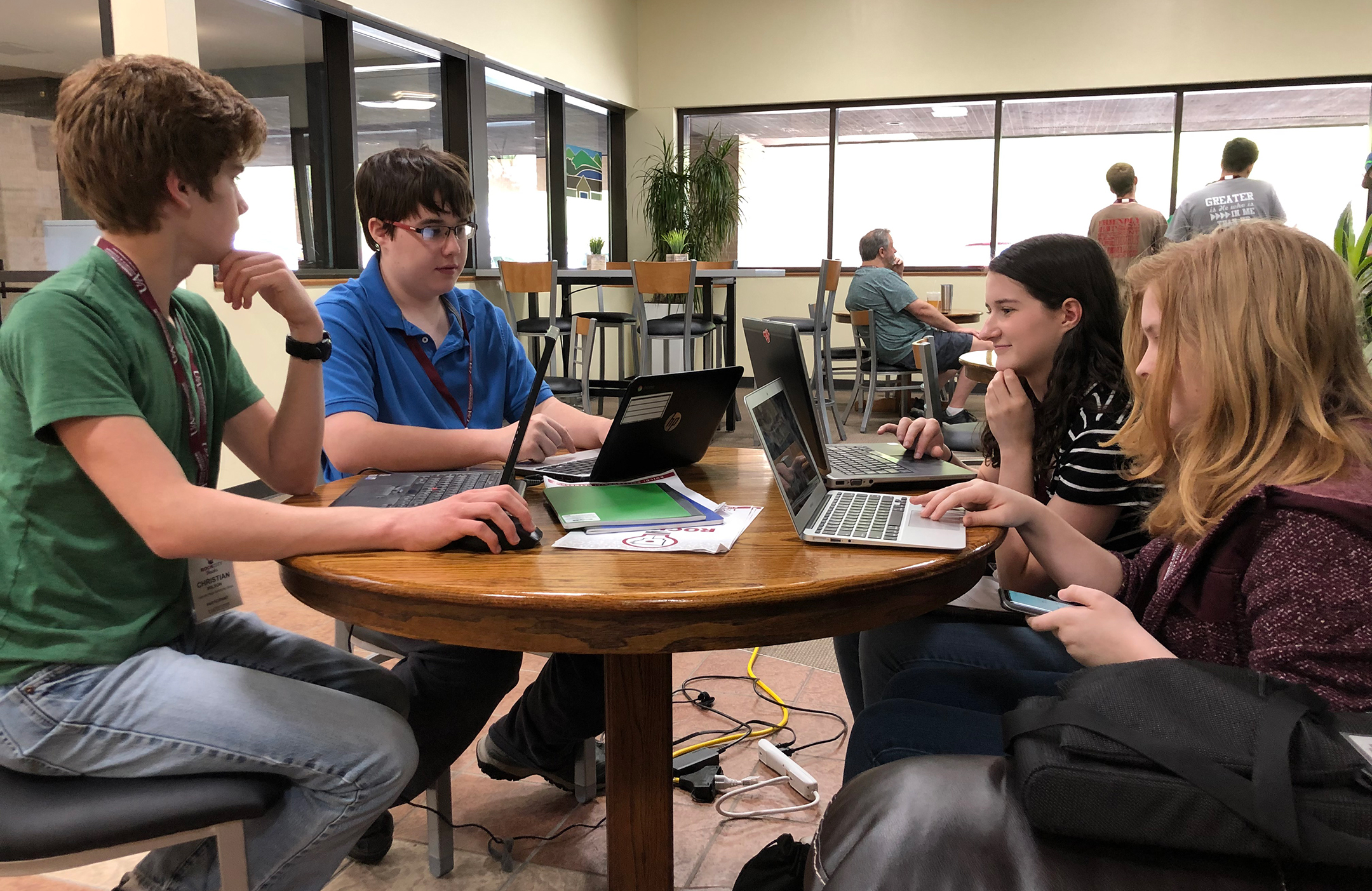 Four high school students, two boys and two girls, sit at a table each with their laptops open at a hackathon