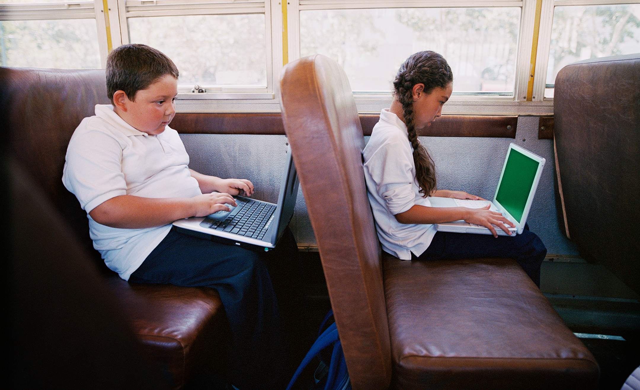 Boy and girl (5-7) on school bus with laptop computer, side view
