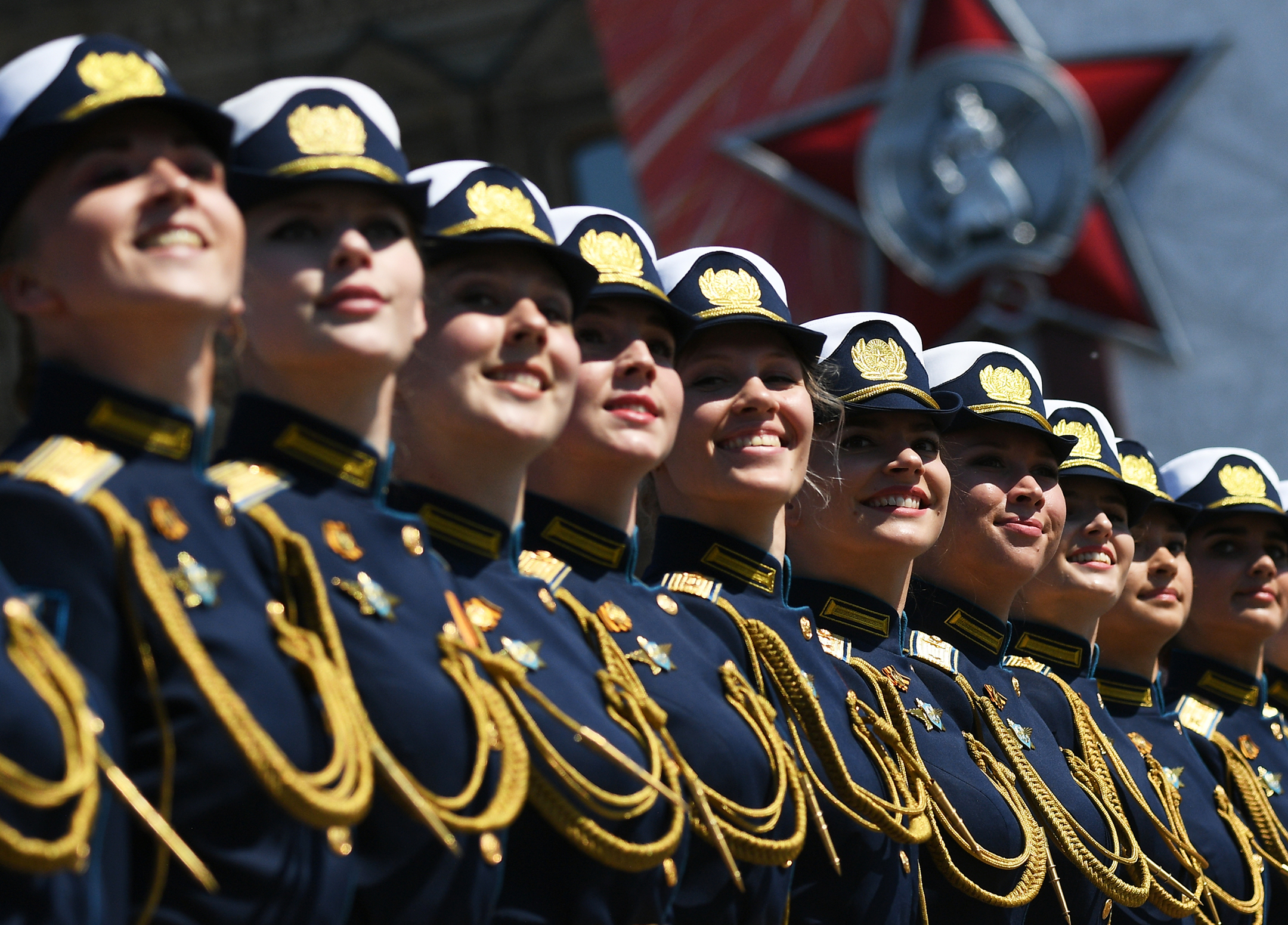 Ten women in military dress uniforms stand in a row with a giant red star in the background