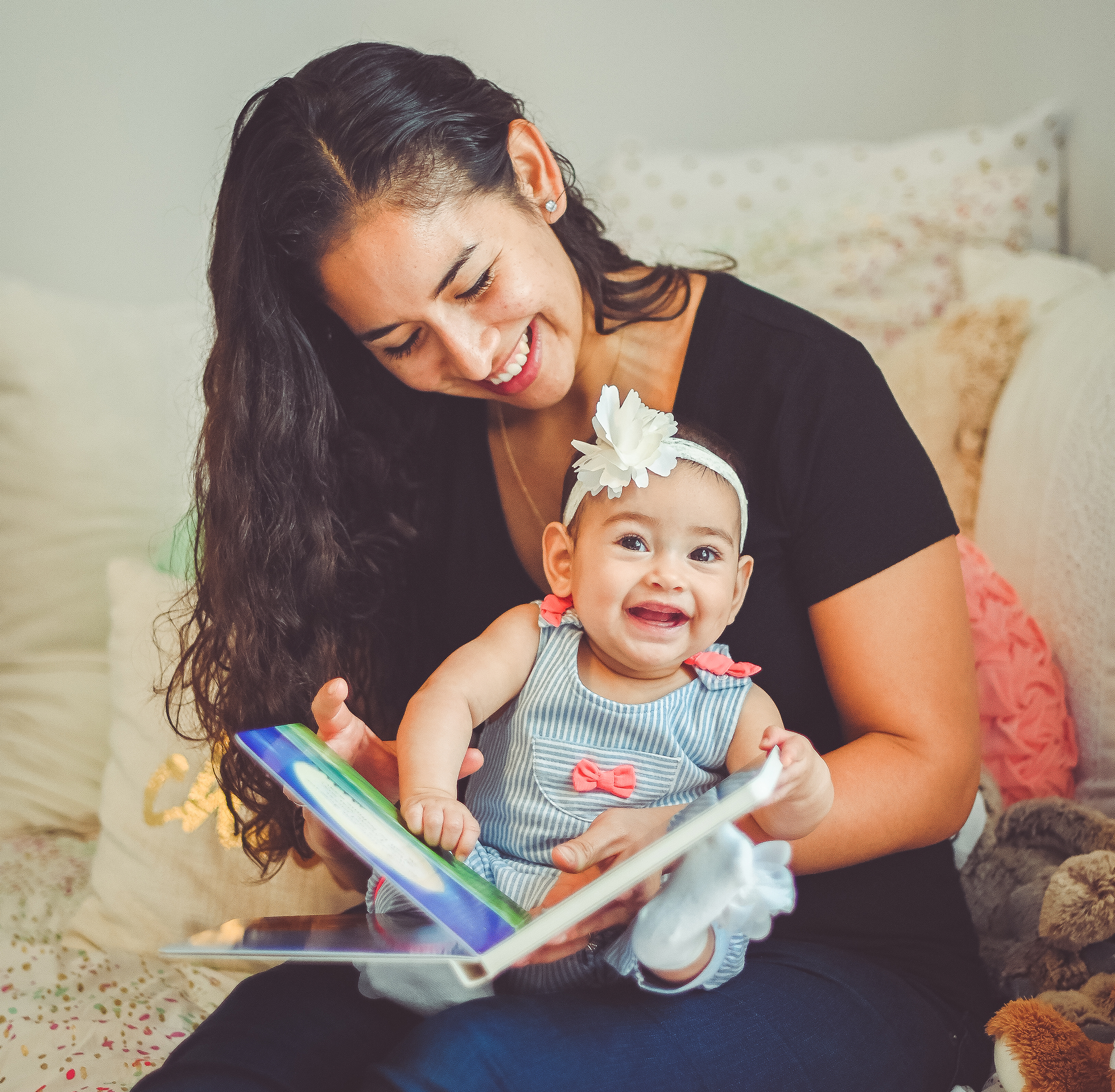 mother and daughter Latina pair read a book together at home in a candid moment. Little girl is around 6 months old and her mother sits on a bed reading with her