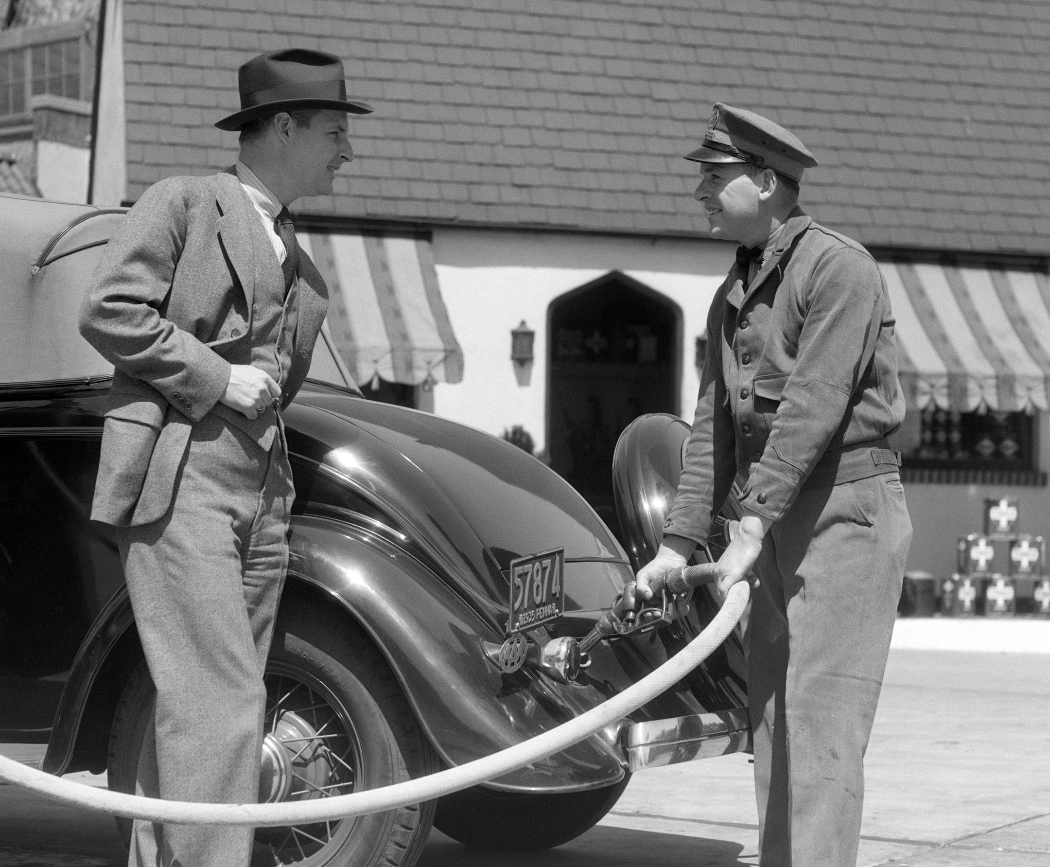 black and white photo circa 1930s of a uniformed Attendant Putting Gasoline In The Tank Of A Coupe While Standing Customer Looks On Smiling 3 piece Suit 