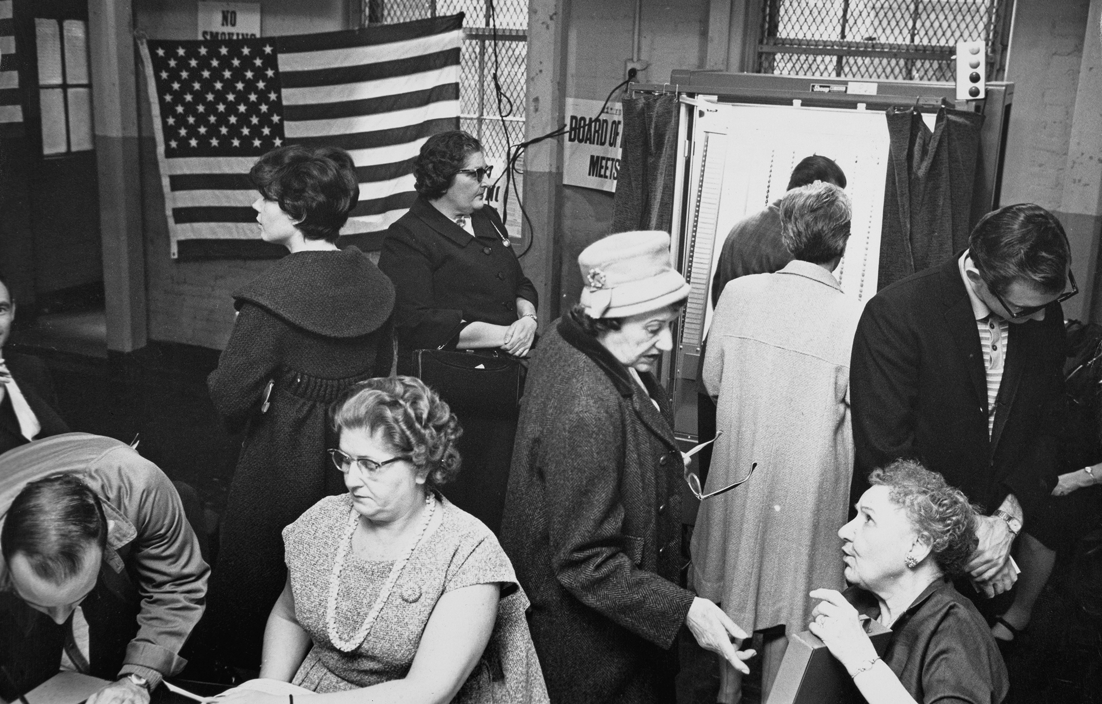 Black and white photo of 9 people, men and women, milling around and voting in a polling place 