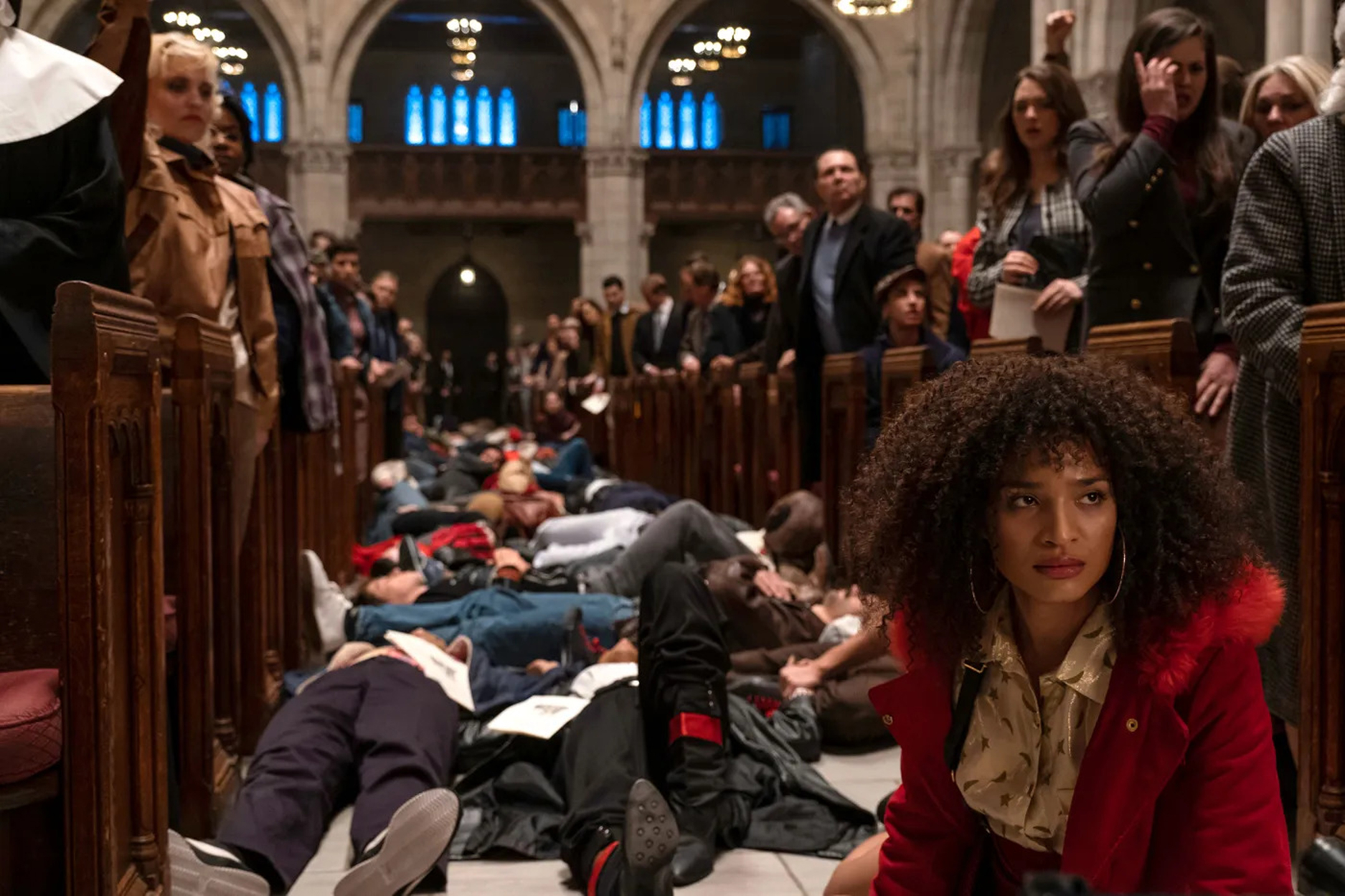 Woman in a red coat sits on the aisle between two rows of church pews, where behind her dozens of people pretend to be dead on the floor while churchgoers in the pews attend mass
