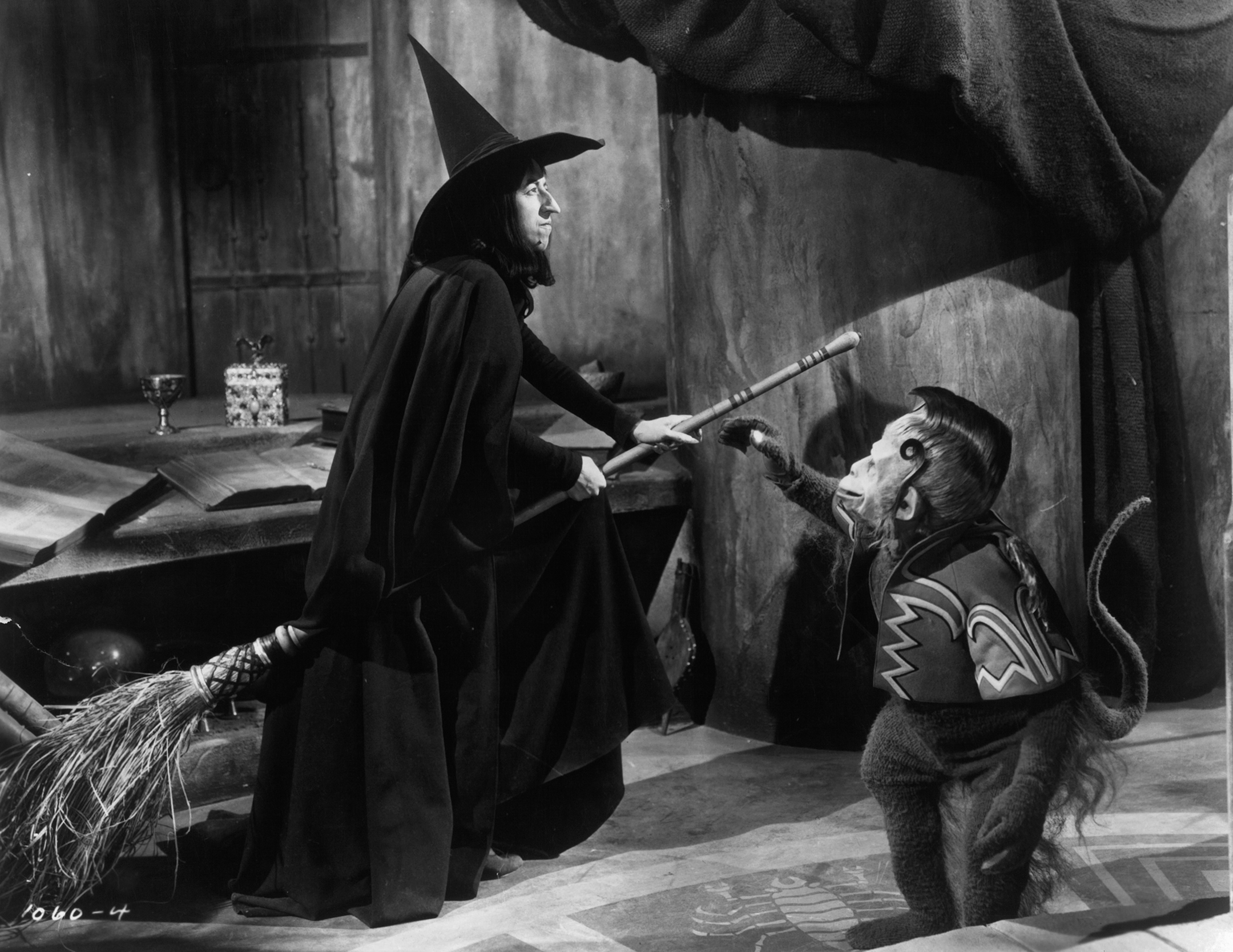 Black and white still from the Wizard of Oz showing the Wicked Witch on her broom with a Flying Monkey character reaching out to her.