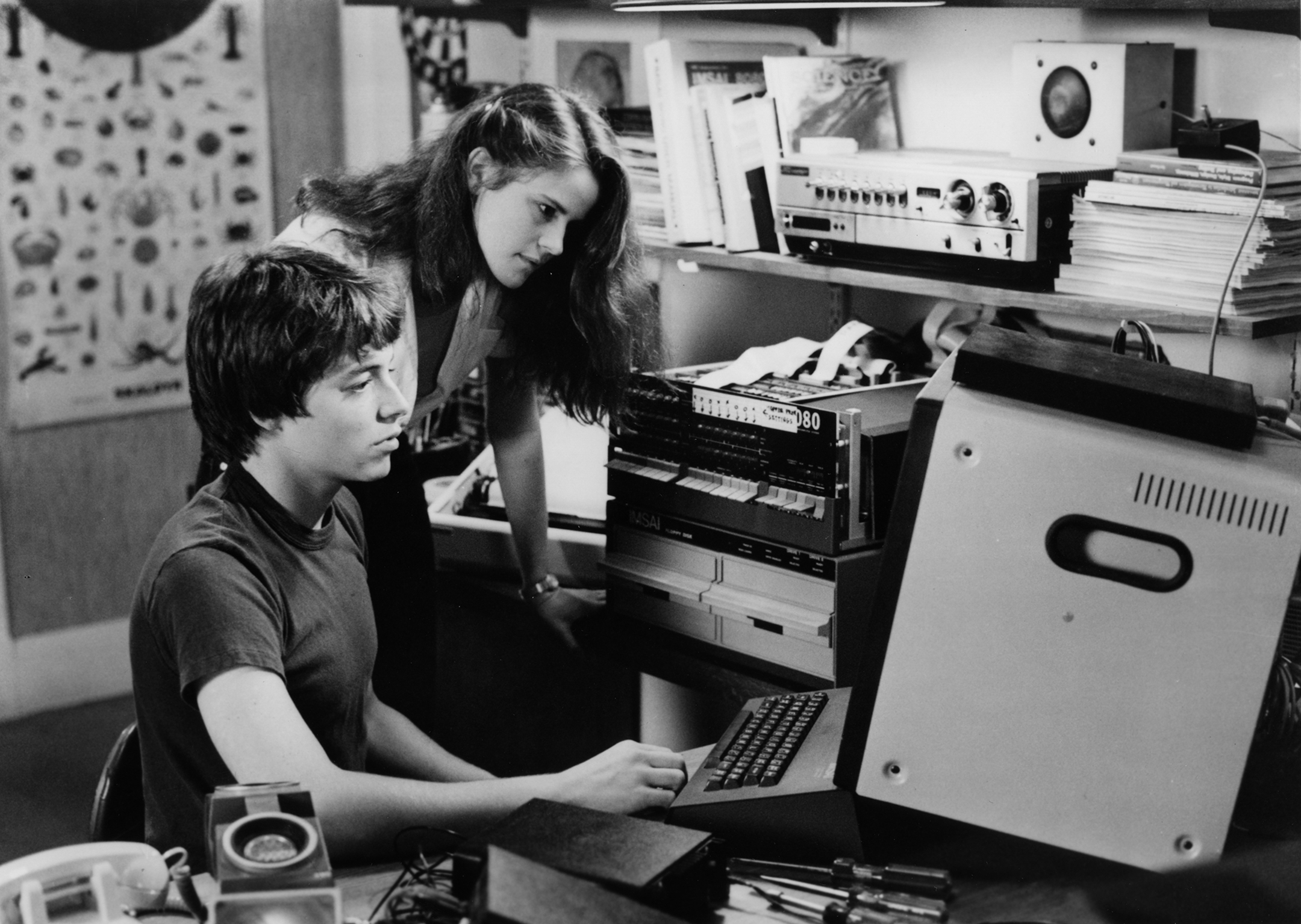 Black and white photo of a young man at a computer in his bedroom and a young woman watching the screen over his shoulder