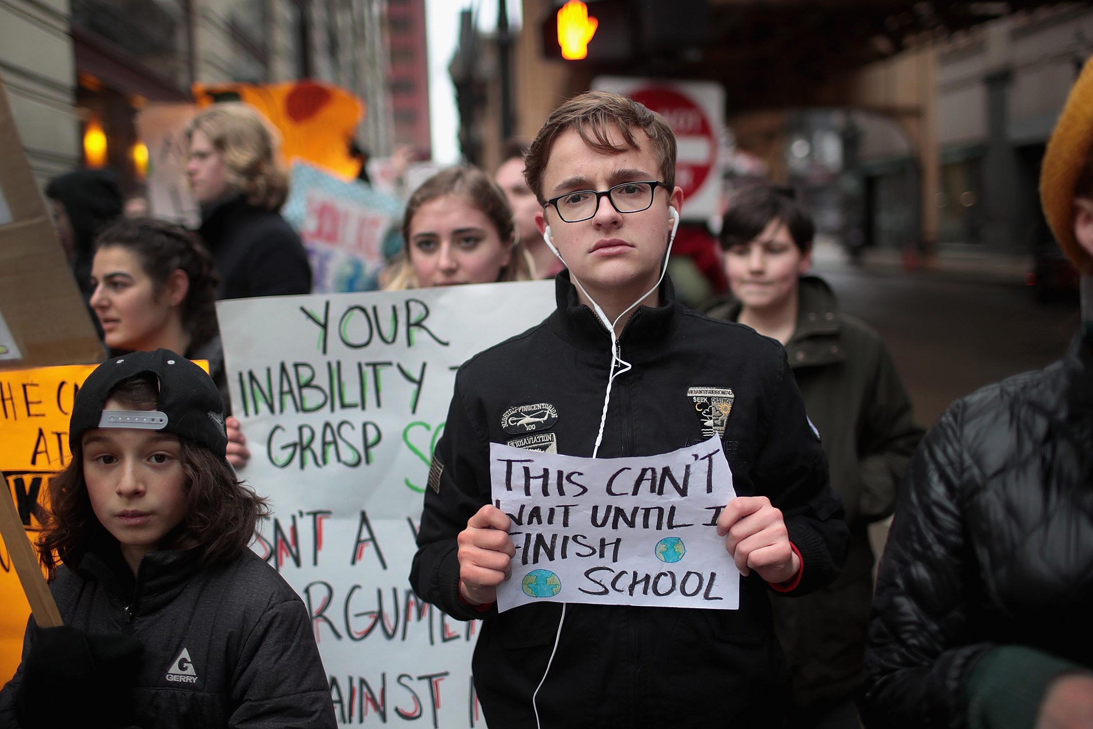 College students at a protest, a young man in the front of the group with glasses holding a sign that reads "This can't wait until I finish school"