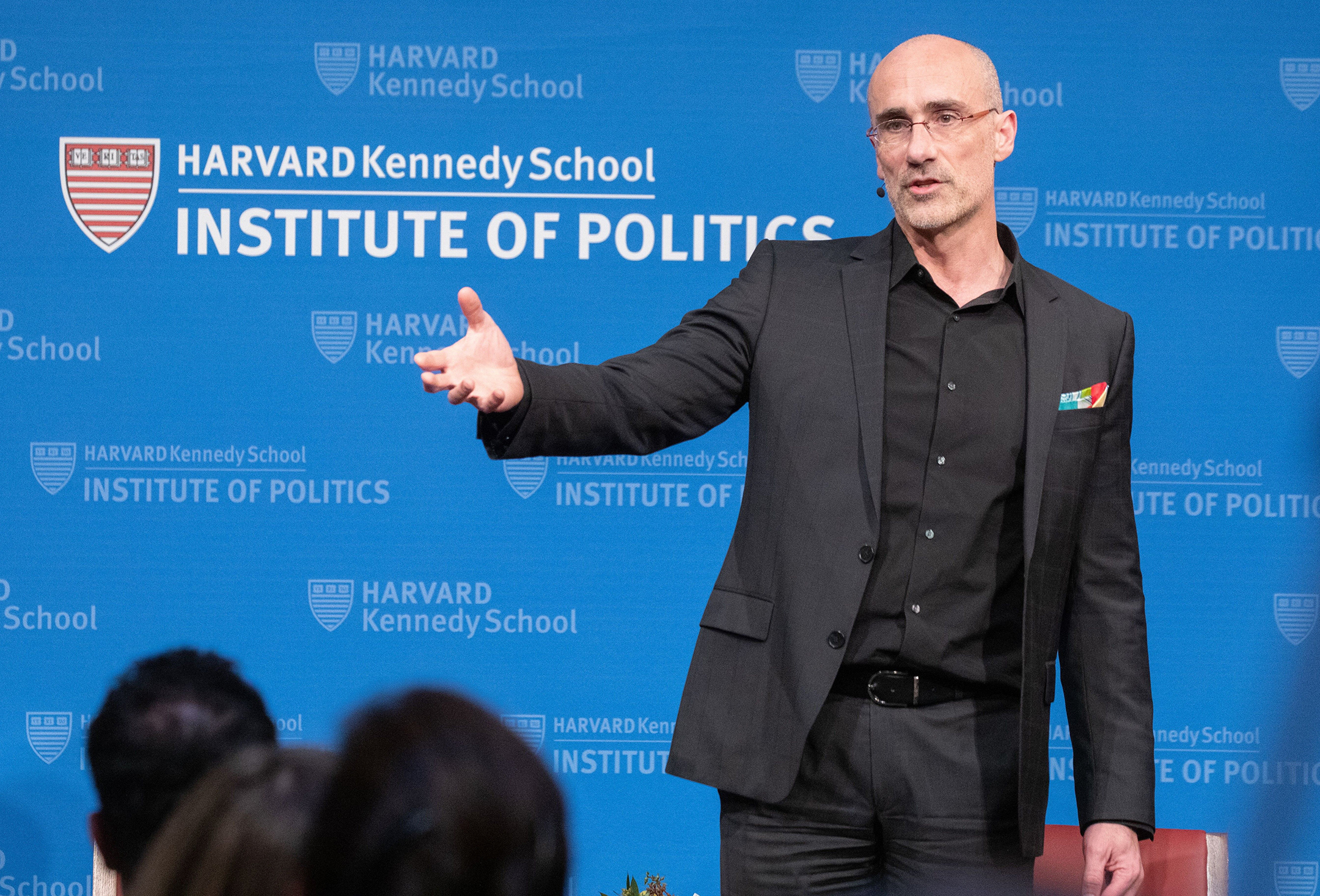 Photo of a man in a black blazer, black button-down shirt, and black pants speaking to a crowd in front of a backdrop that reads Harvard Kennedy School Institute of Politics