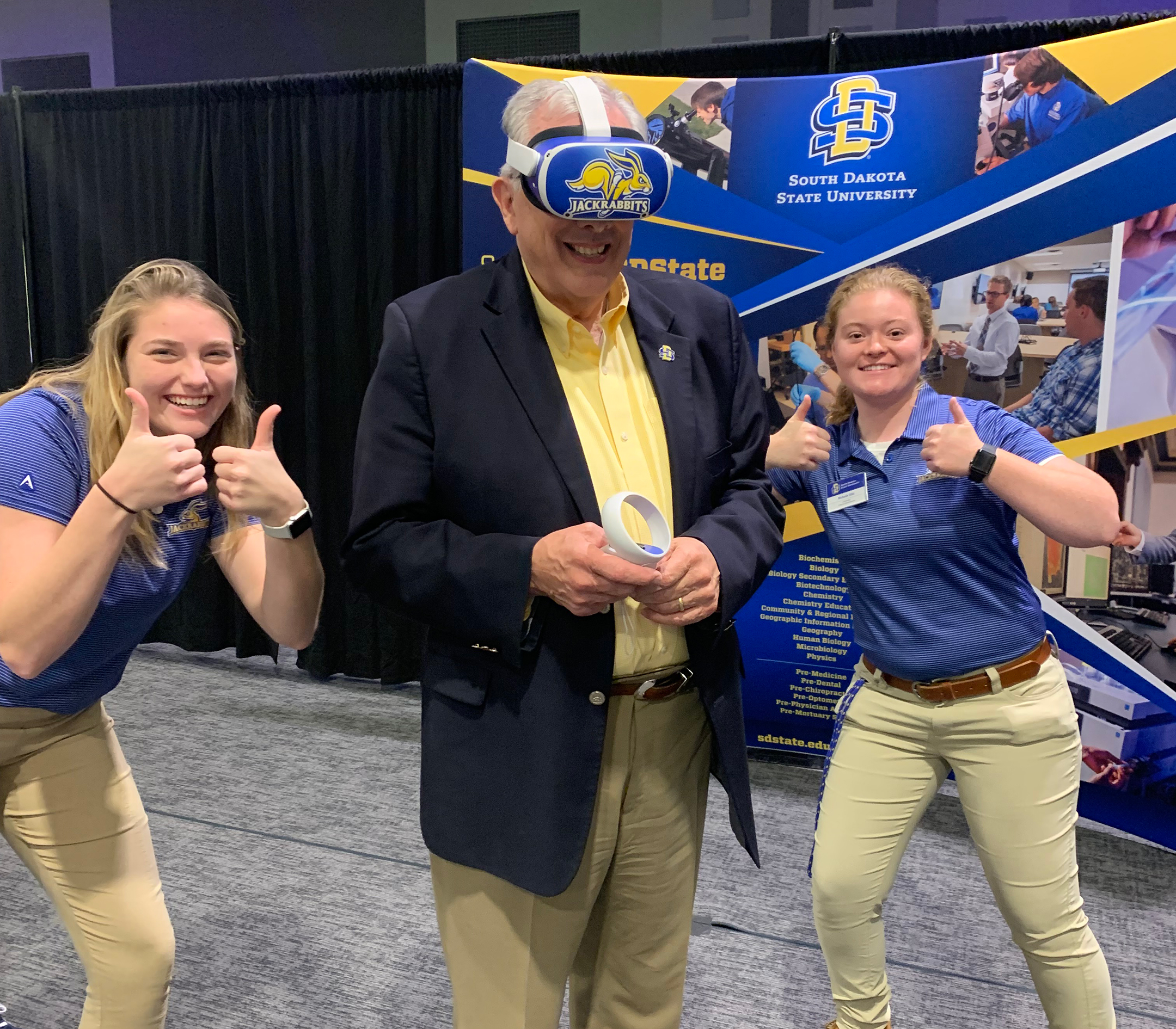 Two smiling young women with thumbs up and an older smiling man wearing a VR headset standing between them