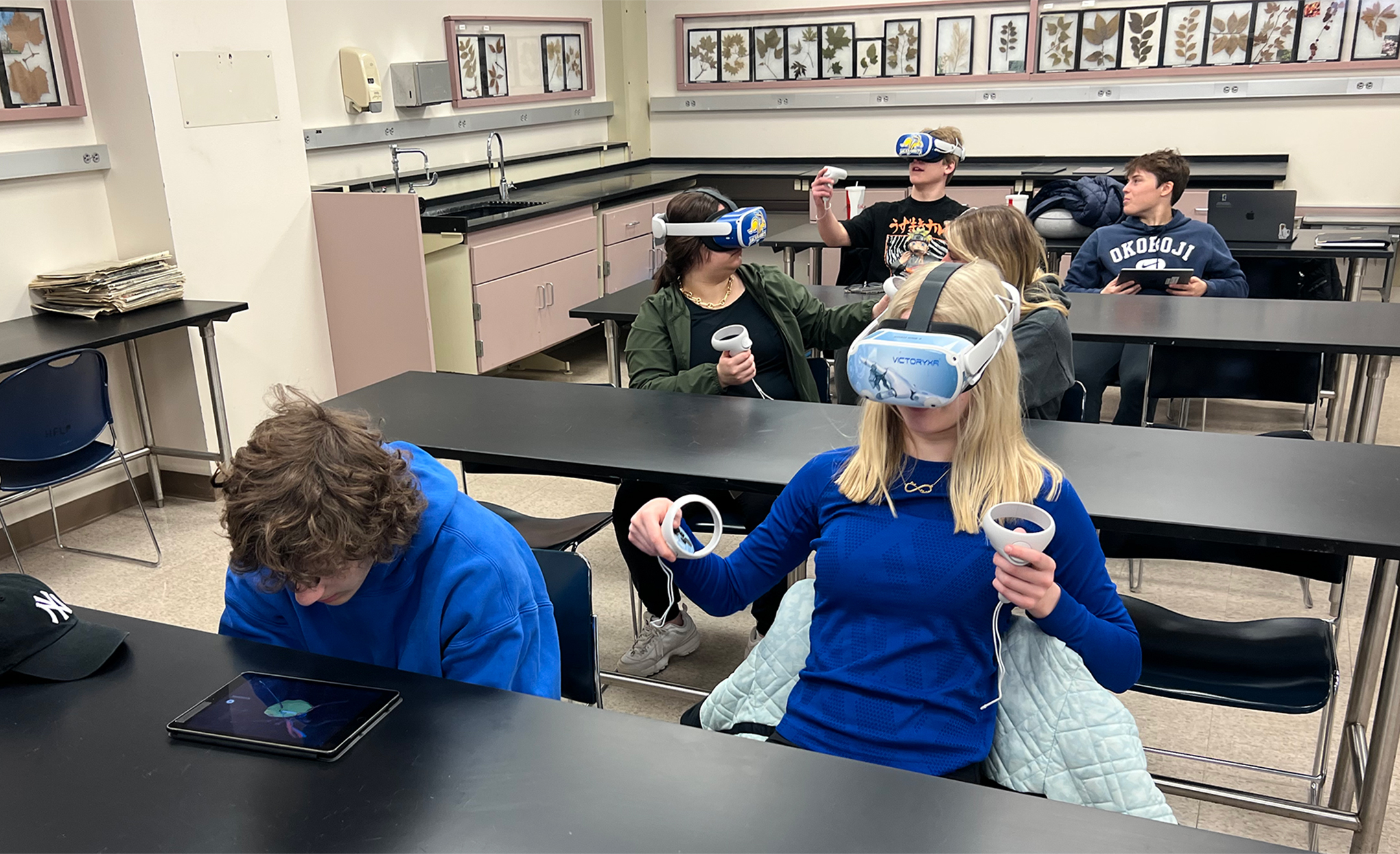 Six high school students sitting at three tables, with one student at each table wearing a VR headset