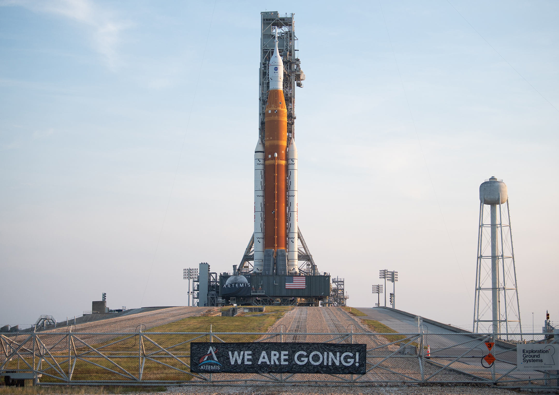 Large booster rocket sits on a mobile mover, behind a fence with a banner that reads We Are Going!