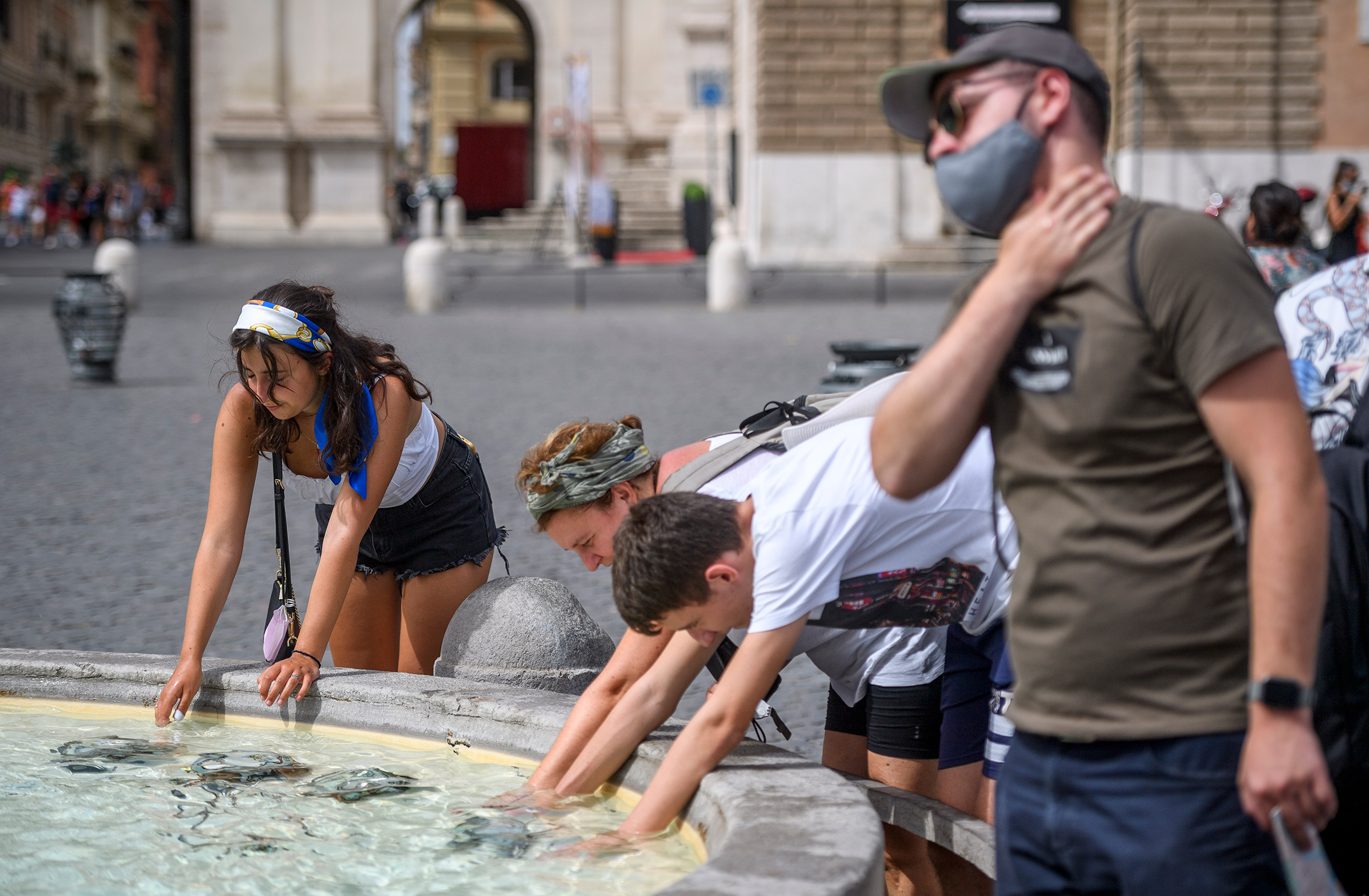 There people, one man and two women, reach their hands into a fountain while a second man slaps water on his neck