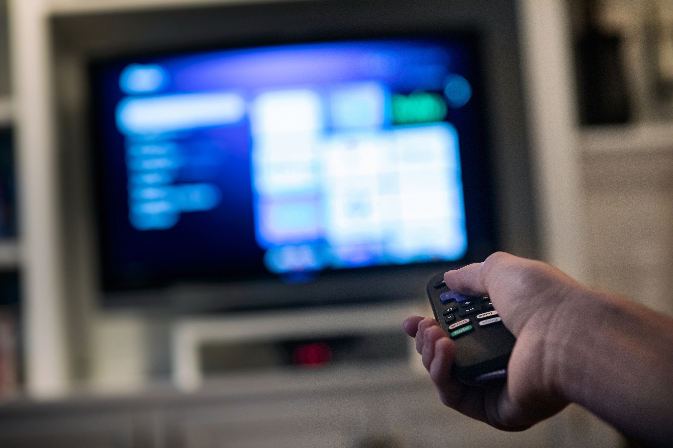 Hand clutching a remote in the foreground, an out-of-focus TV in the background