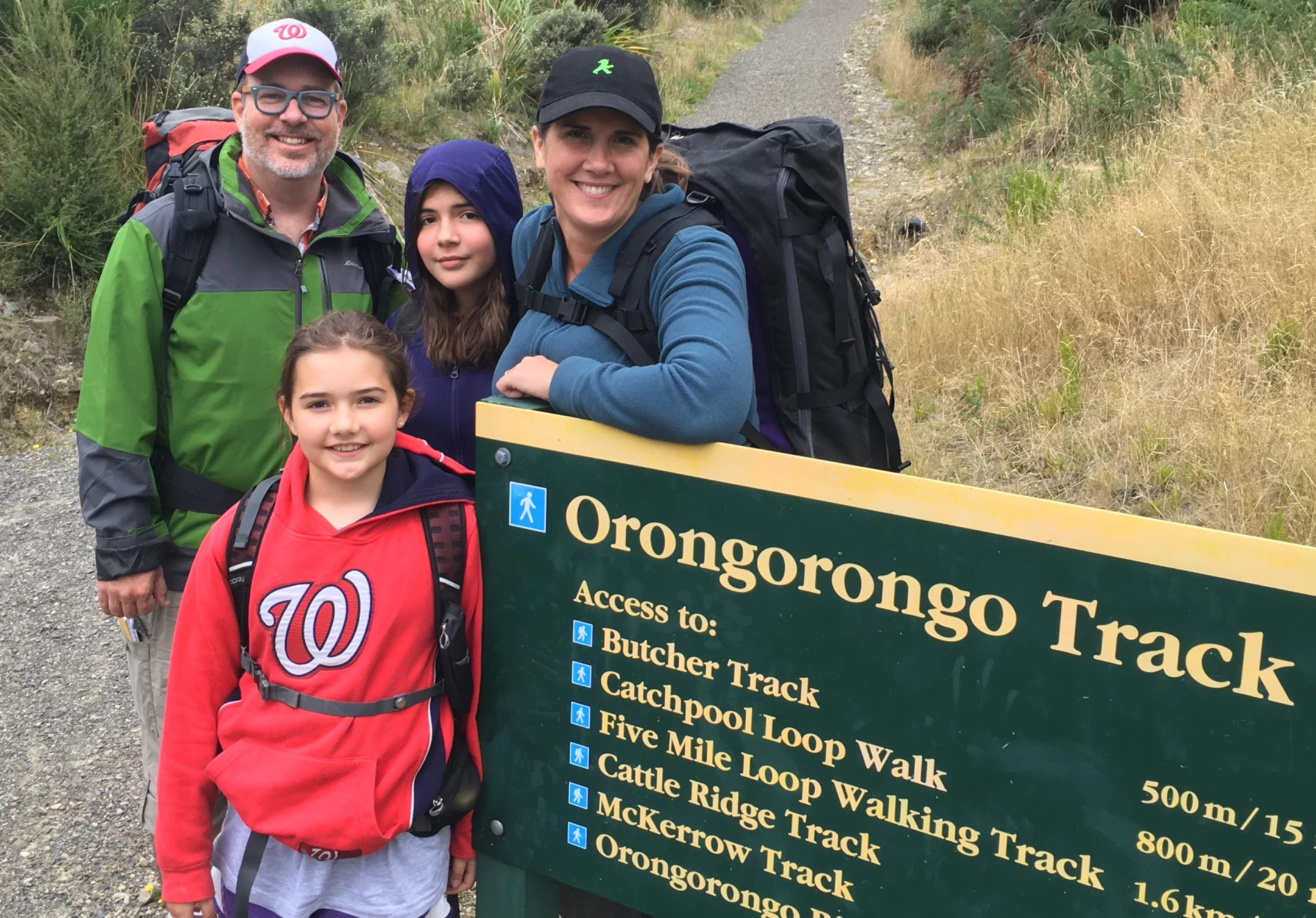 Dan Kois and his wife with their two daughters pose and smile next to a sign that reads Orongorongo Track