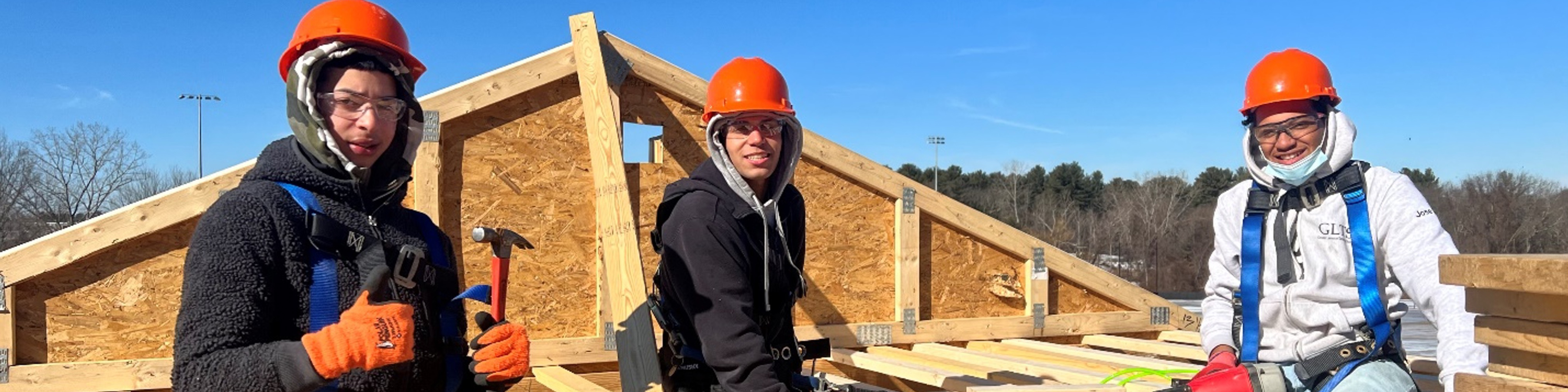 Three high school students in hard hats and coats hold tools and give thumbs up sitting atop wood on a house building project site