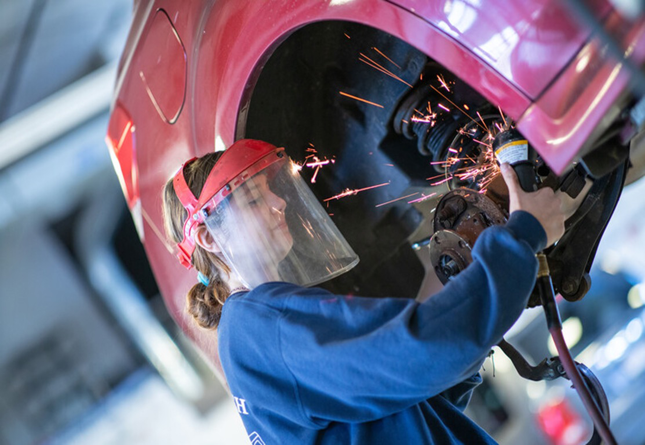 Female high school student wearing a hard hat and plastic face shield using a grinder on a car wheel, creating sparks