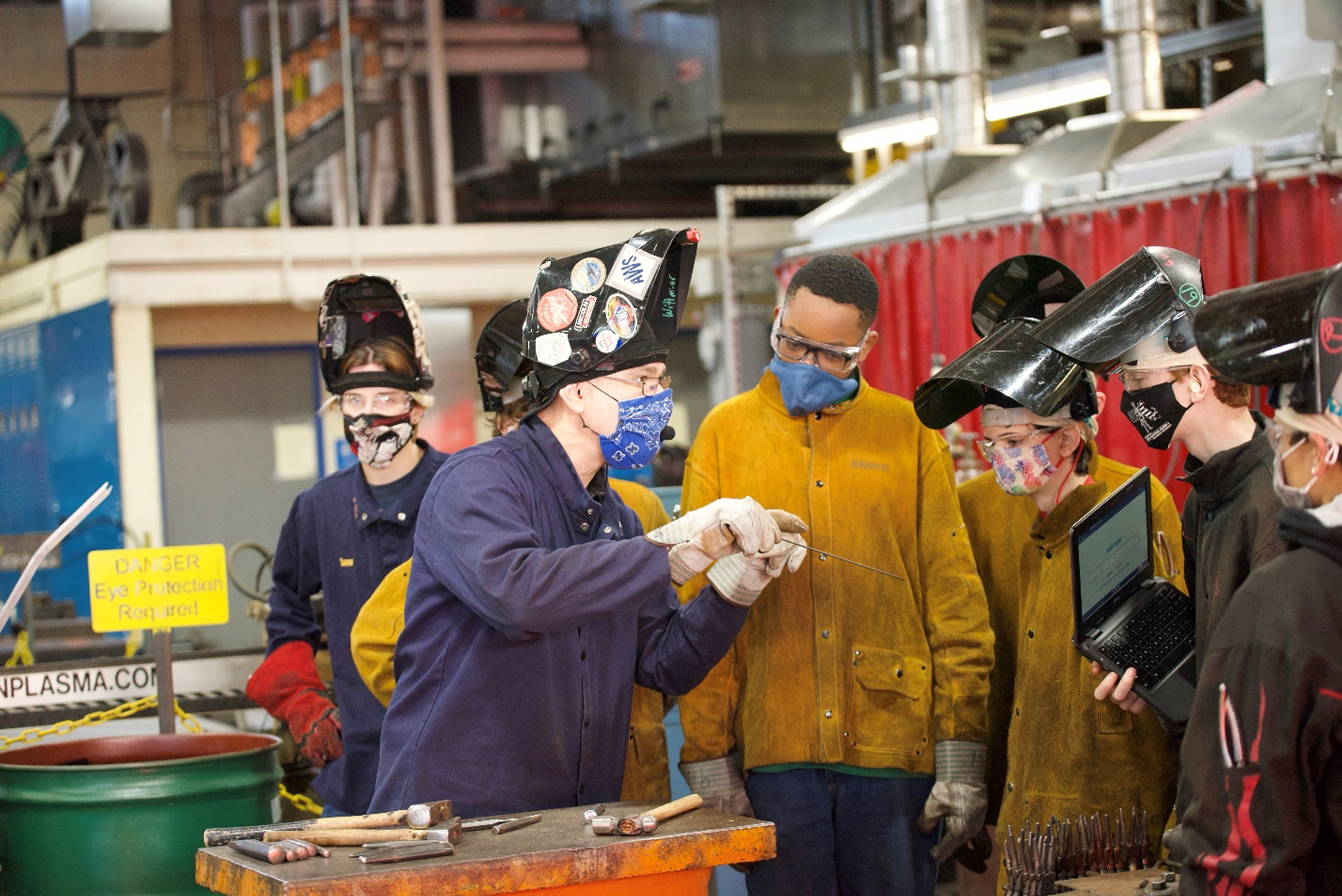 Female teacher in a hard hat and face mask demonstrating a tool to a group of six high school students in a shop class environment 