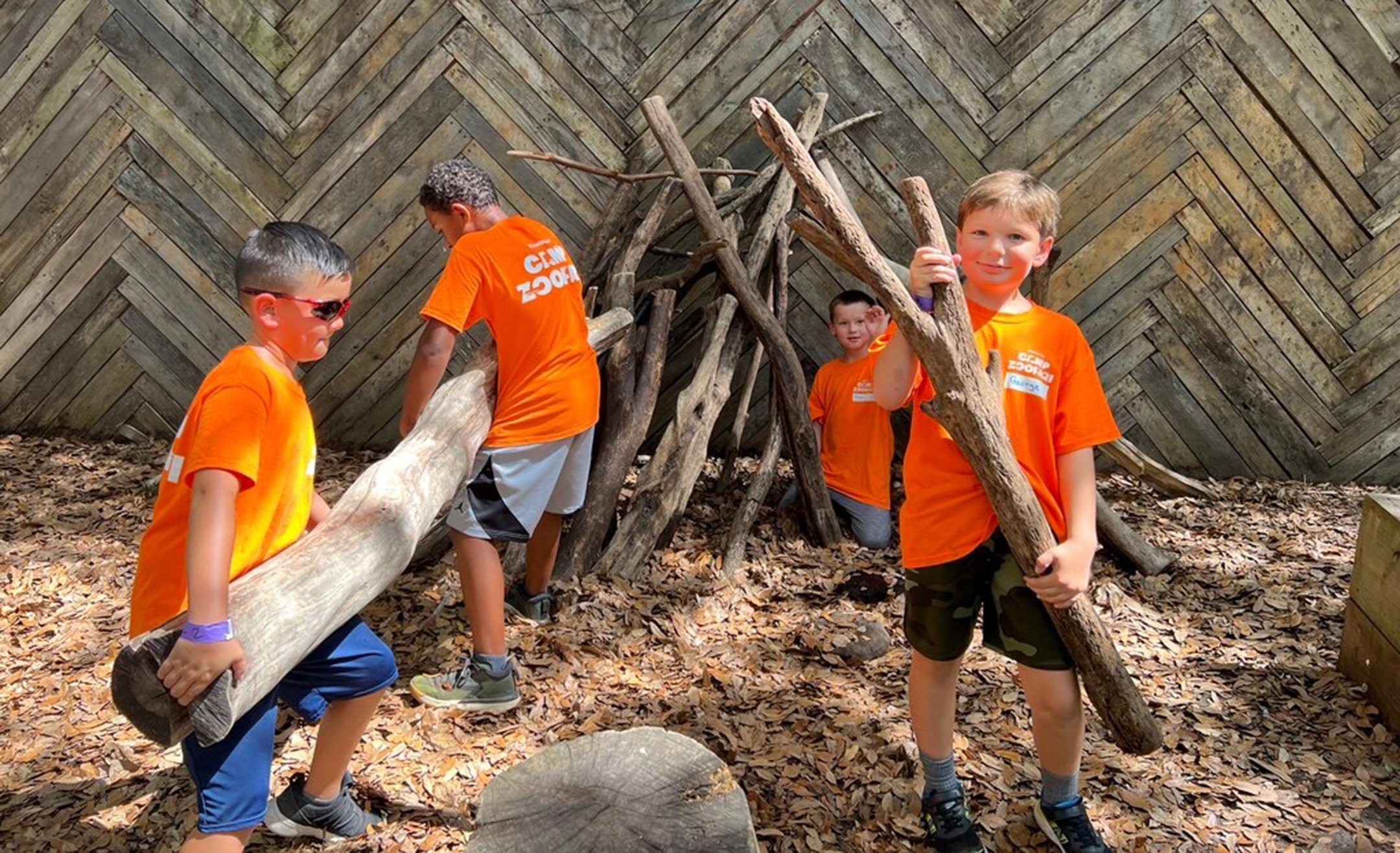 Four boys in orange shirts lift large tree trunks and twigs at an outdoor summer camp