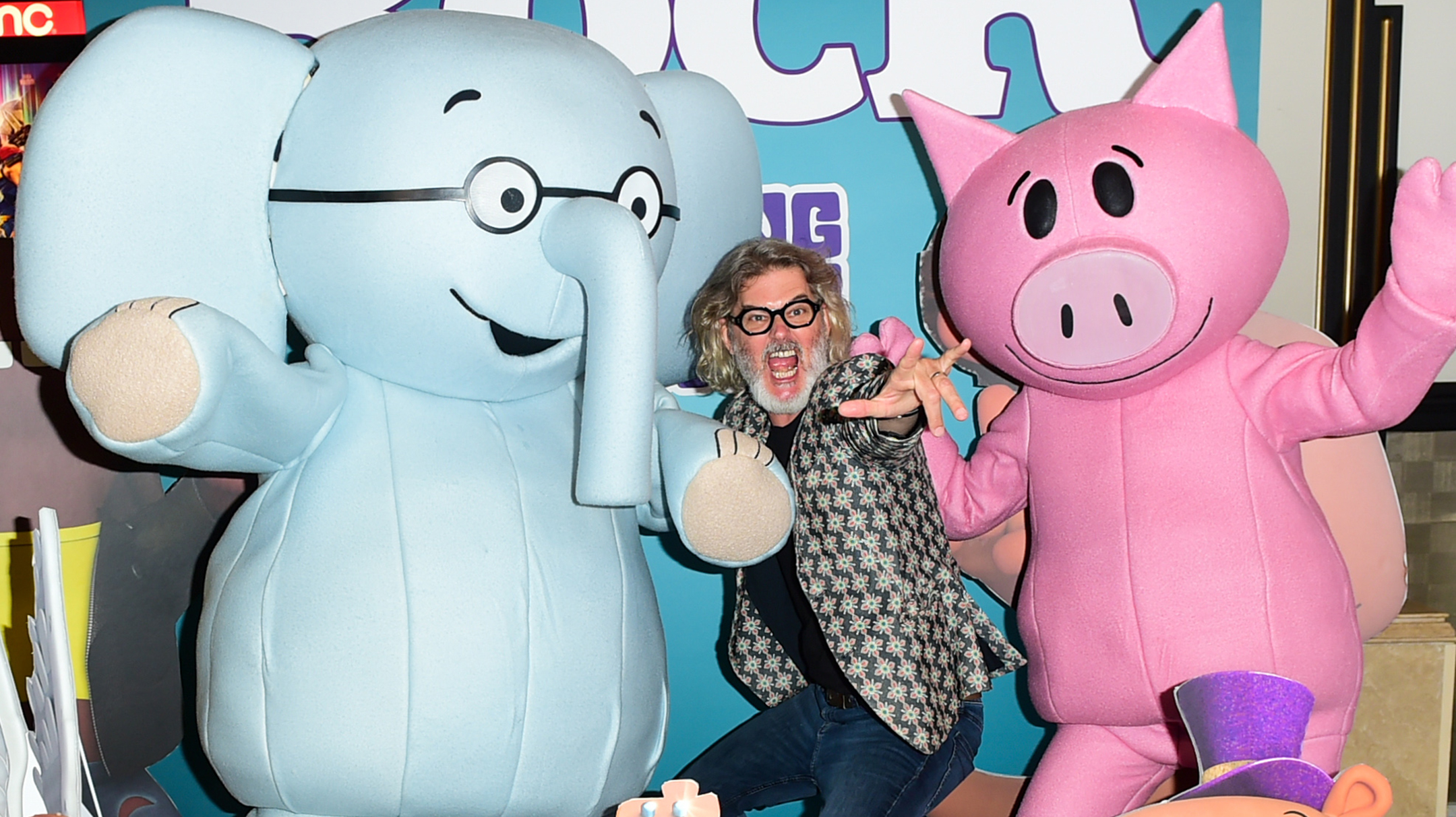 A man in a blazer and black glasses makes a wild expression while being flanked by costumed characters of an elephant on the left and a pig on the right