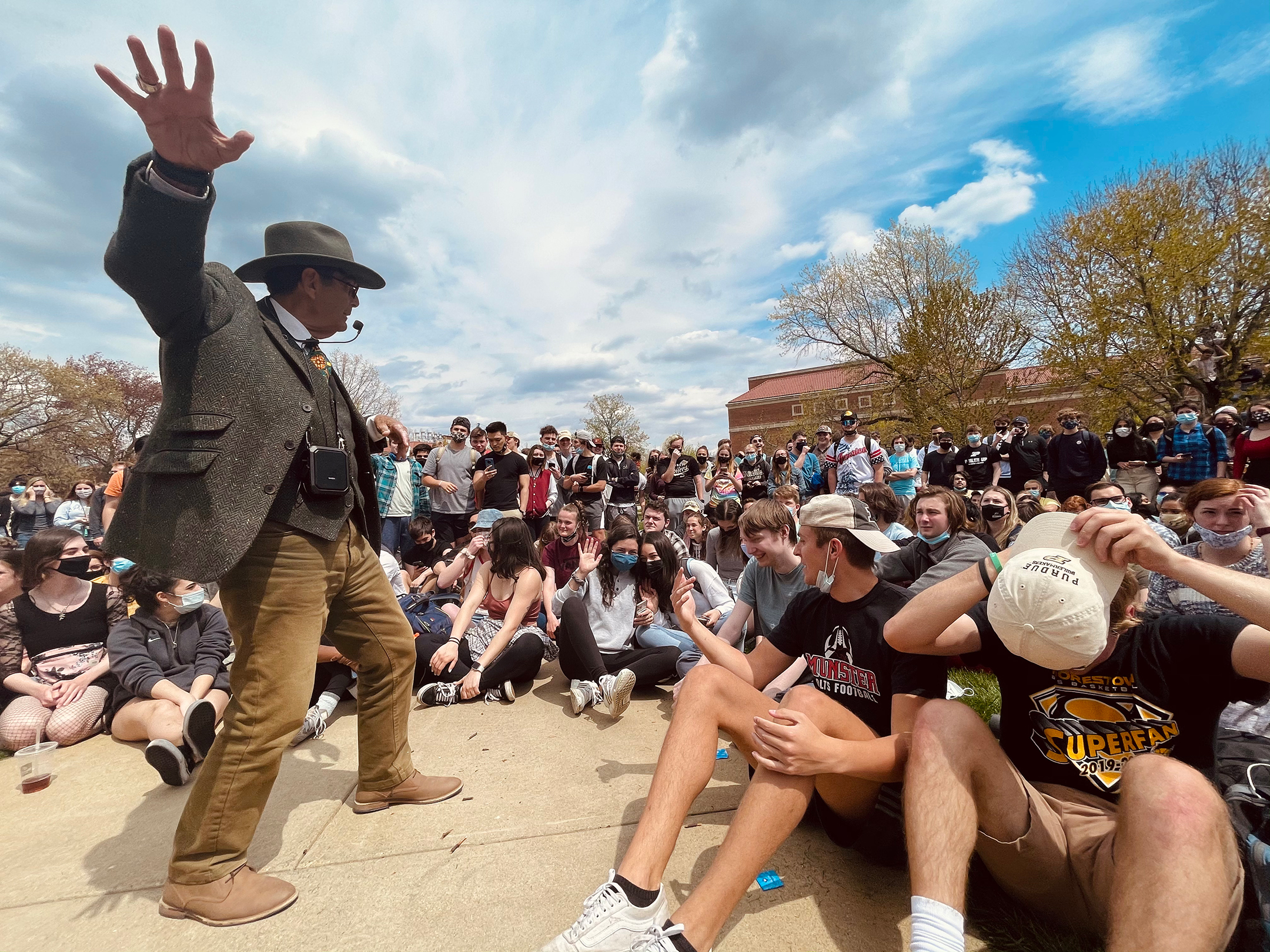 Man with outstretched arm and wearing a microphone in front of a large outdoor crowd of college students