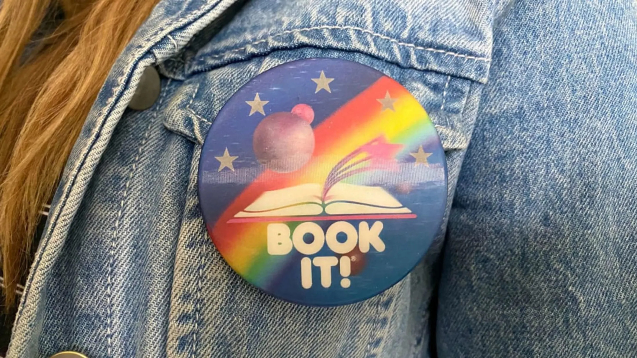 Close up of a pin that reads Book It on a denim jacket worn by a young woman