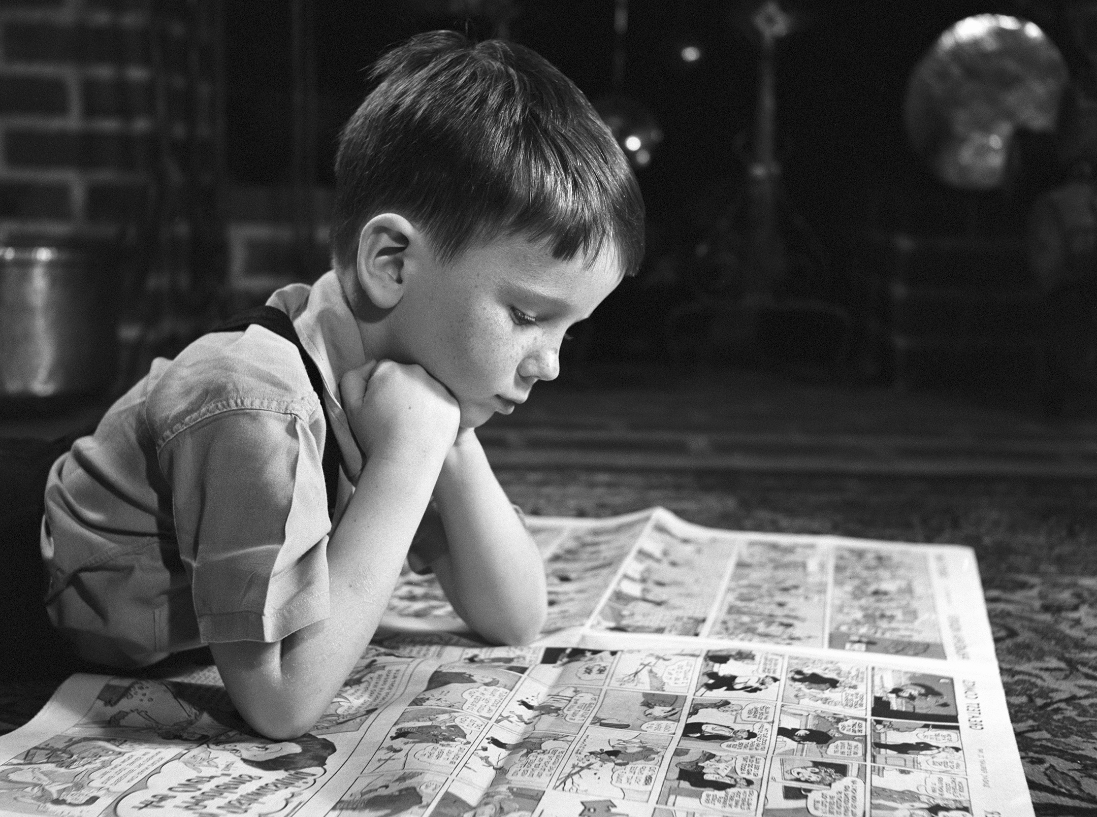 Black and white photo of a boy reading comic newspaper pages on the floor inside a house.